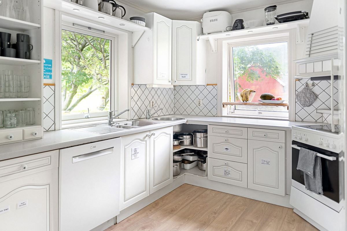 Sunlit white corner kitchen with sink, dishwasher, oven, open shelves, tiled backsplash, and windows with greenery.