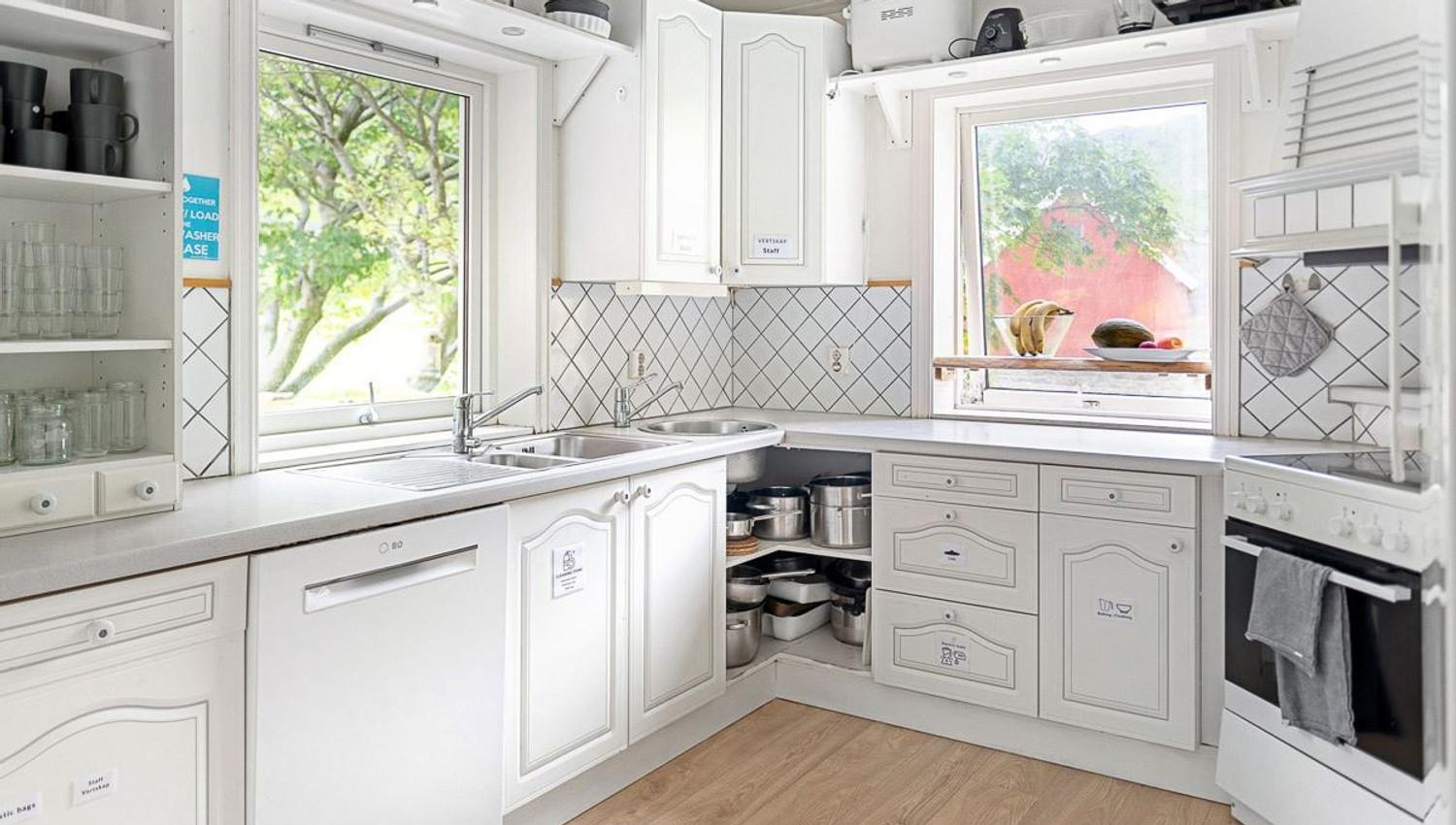 Sunlit white corner kitchen with sink, dishwasher, oven, open shelves, tiled backsplash, and windows with greenery.