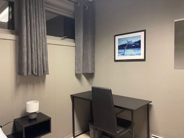 Small room corner with gray curtains, a black desk and chair, a side table lamp, and framed wall art.