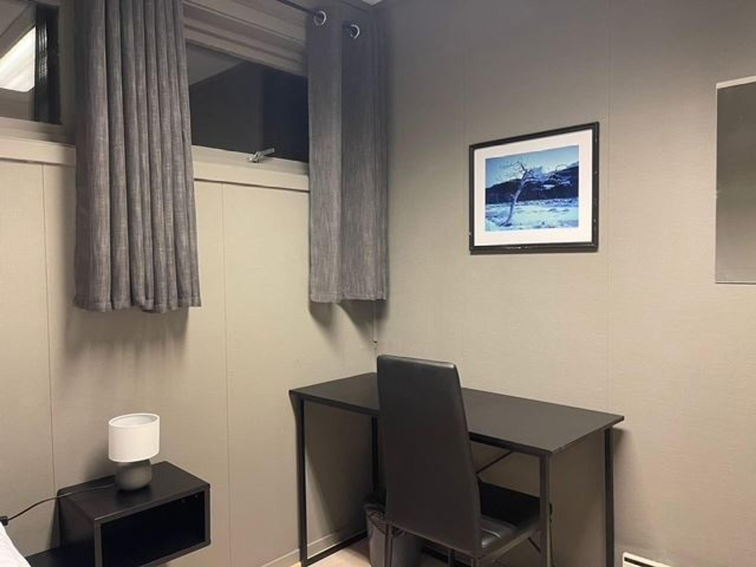 Small room corner with gray curtains, a black desk and chair, a side table lamp, and framed wall art.