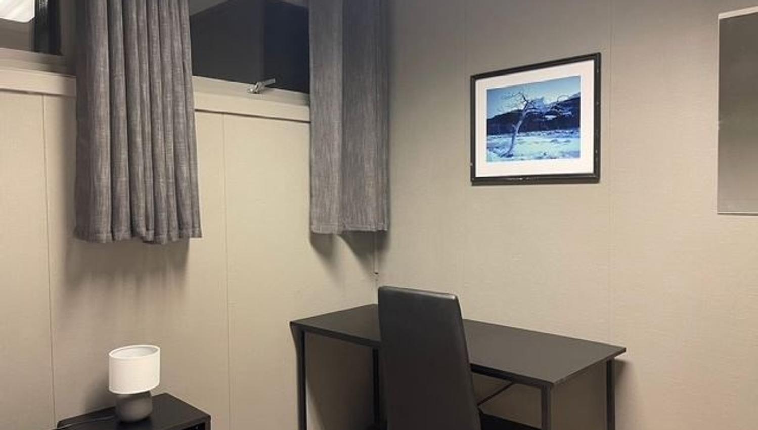 Small room corner with gray curtains, a black desk and chair, a side table lamp, and framed wall art.