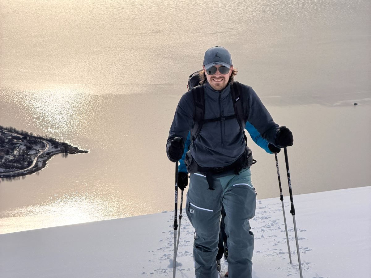 Smiling skiier in winter clothing uses trekking poles on a snowy ridge with a sunlit lake and shoreline below..