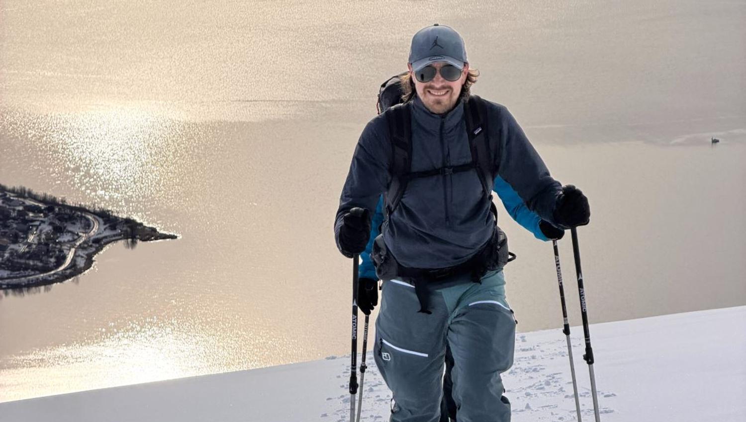 Smiling skiier in winter clothing uses trekking poles on a snowy ridge with a sunlit lake and shoreline below..