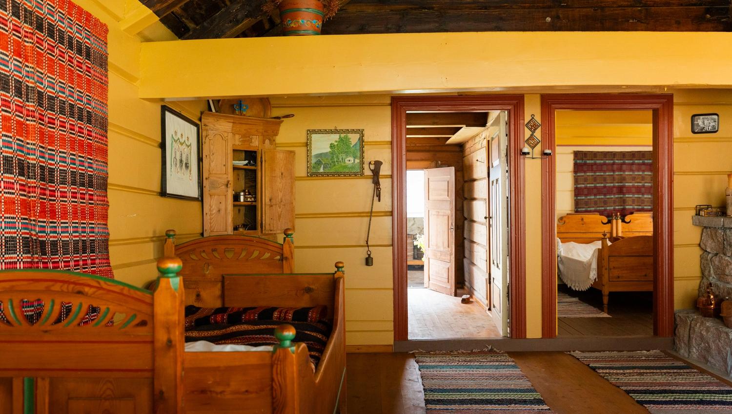 Rustic cabin room with wooden bed frames, striped textiles, wall art, rugs, and open doors to a bedroom.