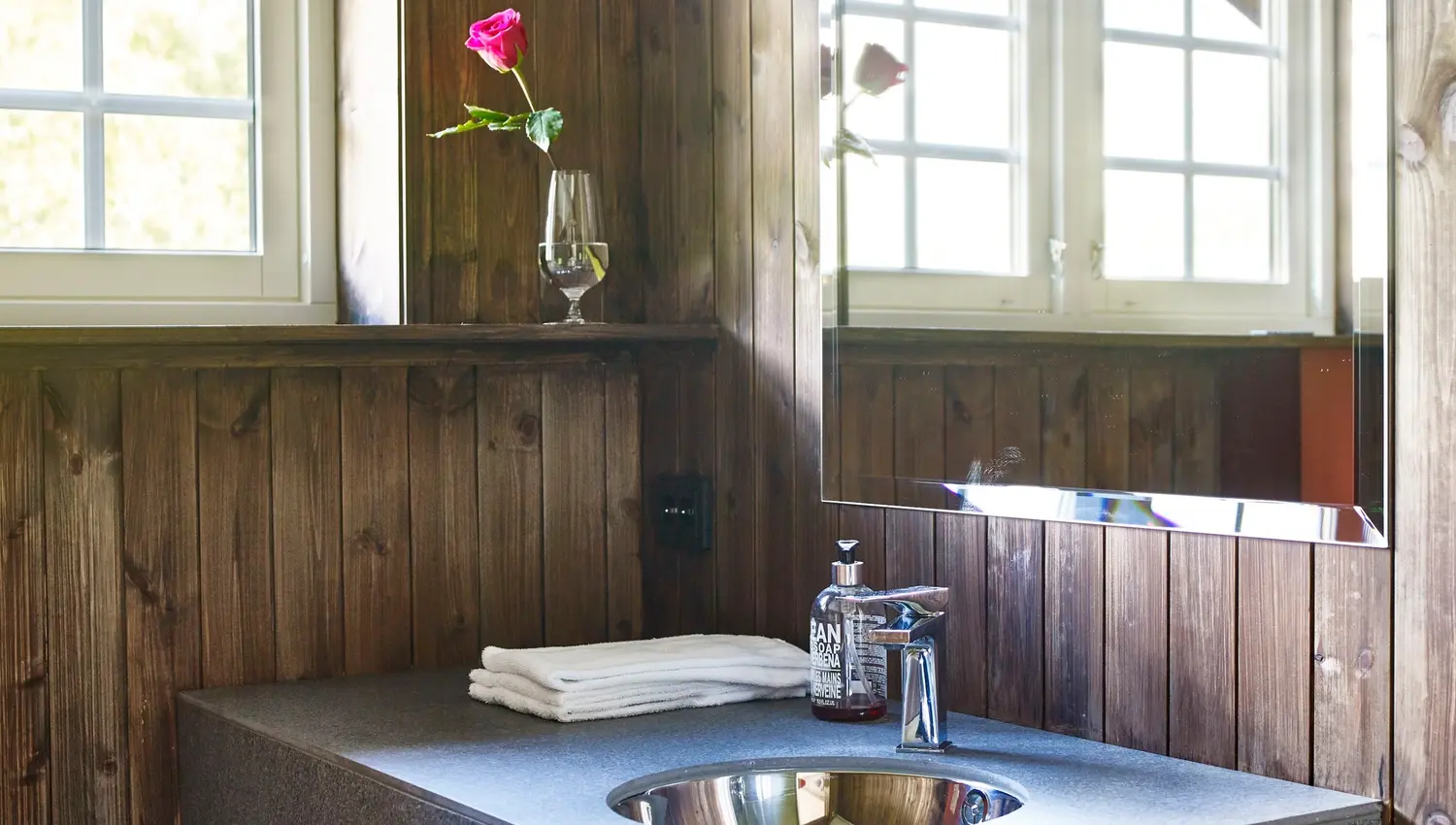 Rustikk baderom med trepanel, vegghengt servant med kran, spegel med lys, vindauge, handkle og rose i glas..