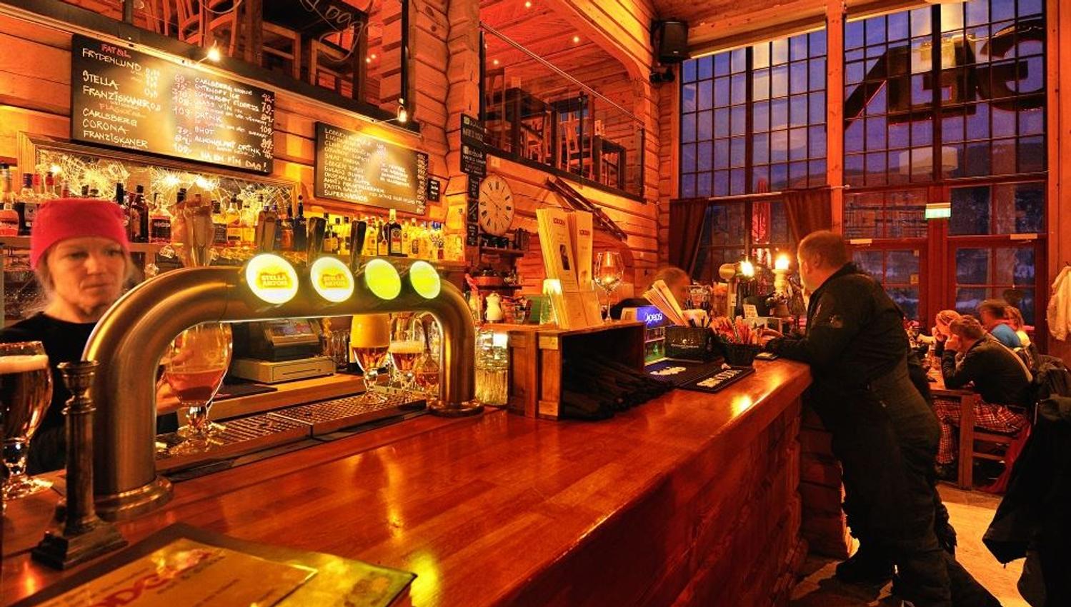 Varmt opplyst bar i treinteriør med ølkraner og flasker, kunder ved disken og store vinduer mot kveld..