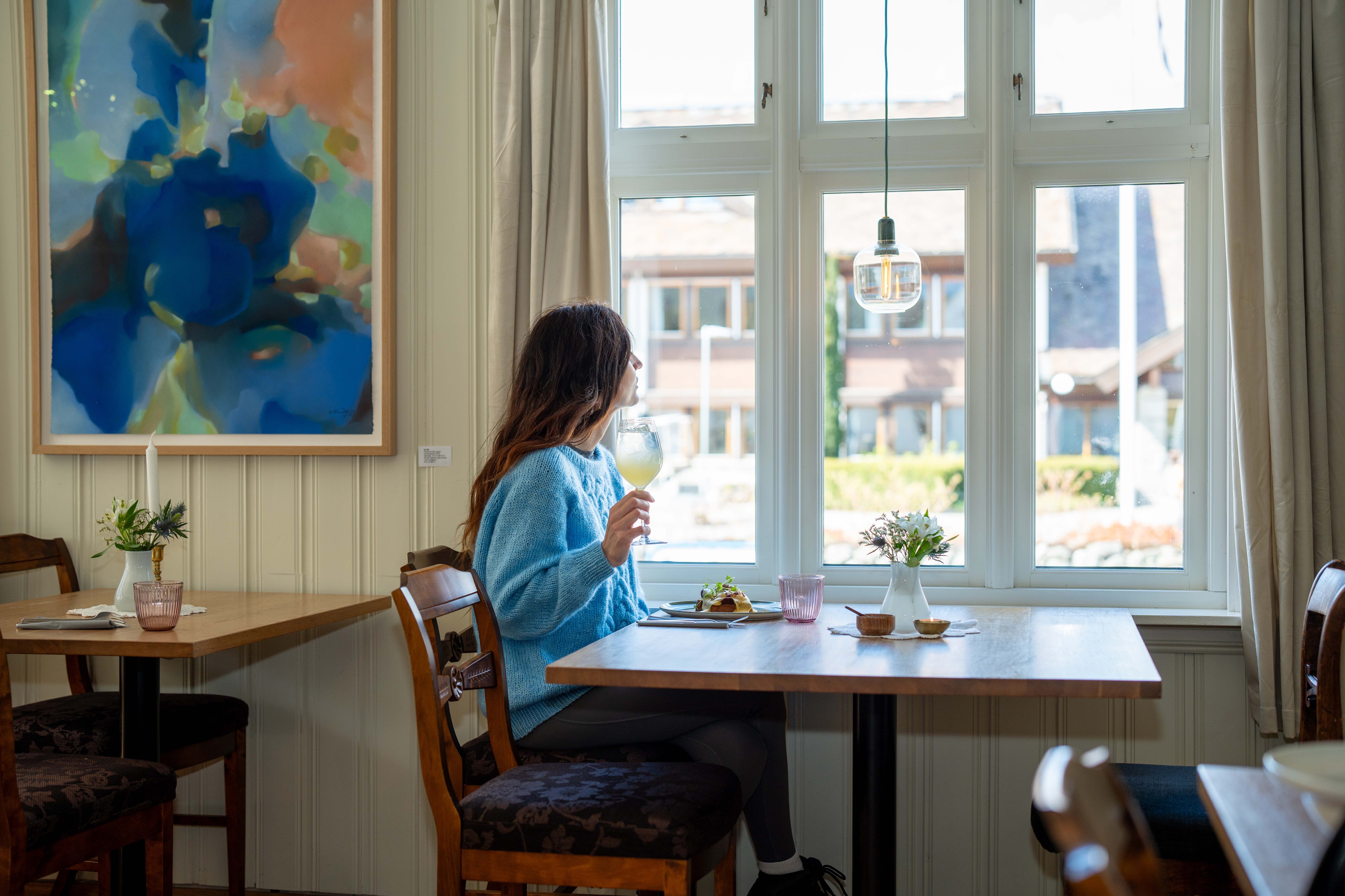 A woman sitting at a table, looking out the window with a glass of juice in her hand.