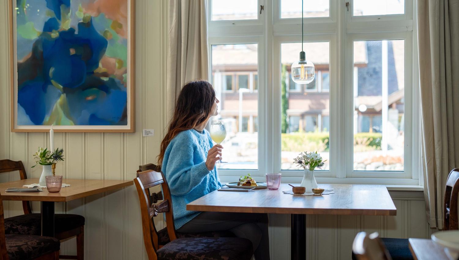 A woman sitting at a table, looking out the window with a glass of juice in her hand.