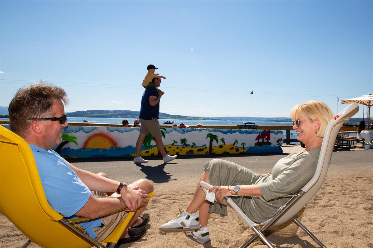 To personer sitter i solstoler på stranden mens en person passerer forbi i bakgrunnen.