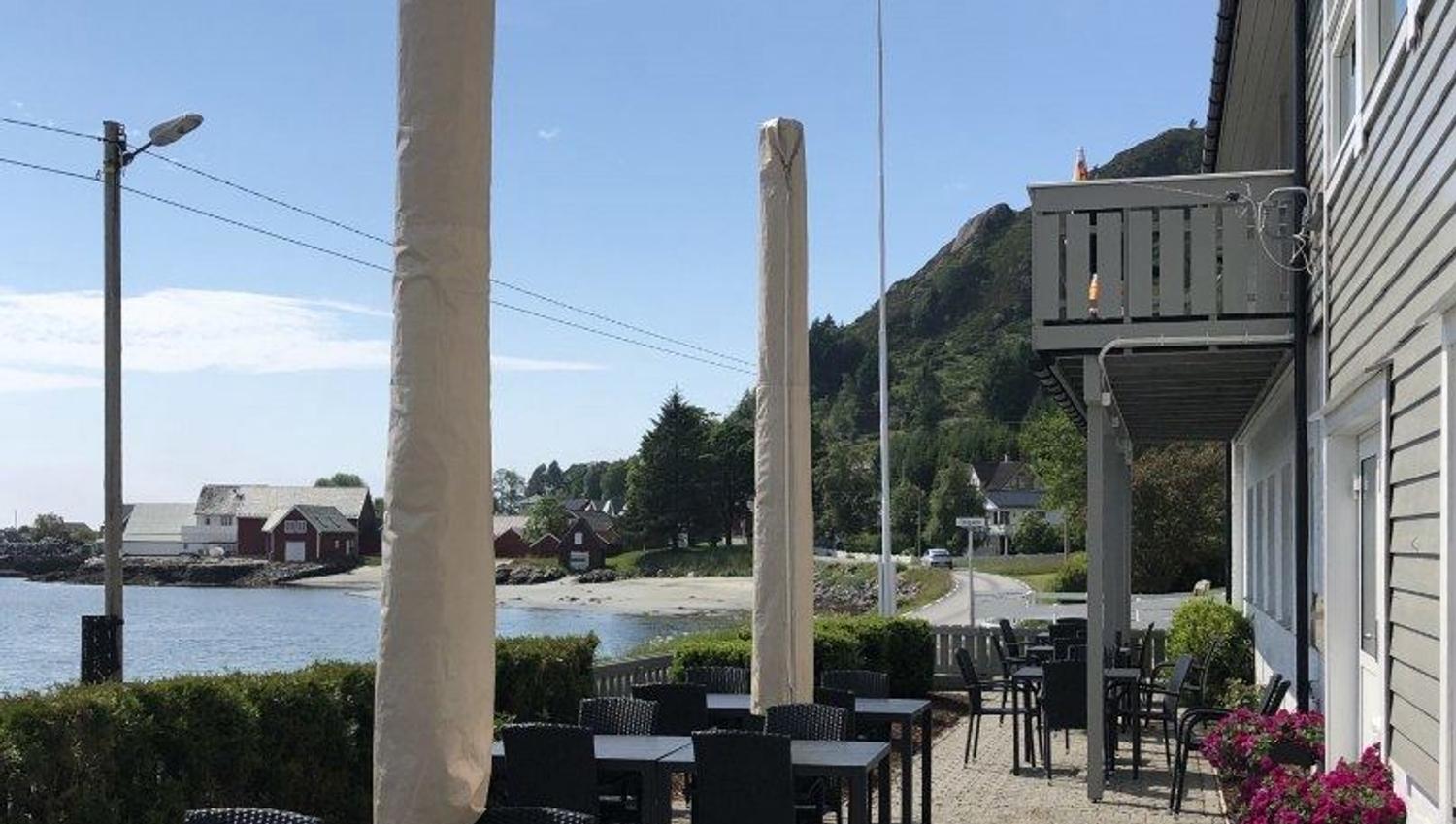 Outdoor cafe patio with tables, closed umbrellas, and a flag beside a waterfront road and hillside houses..