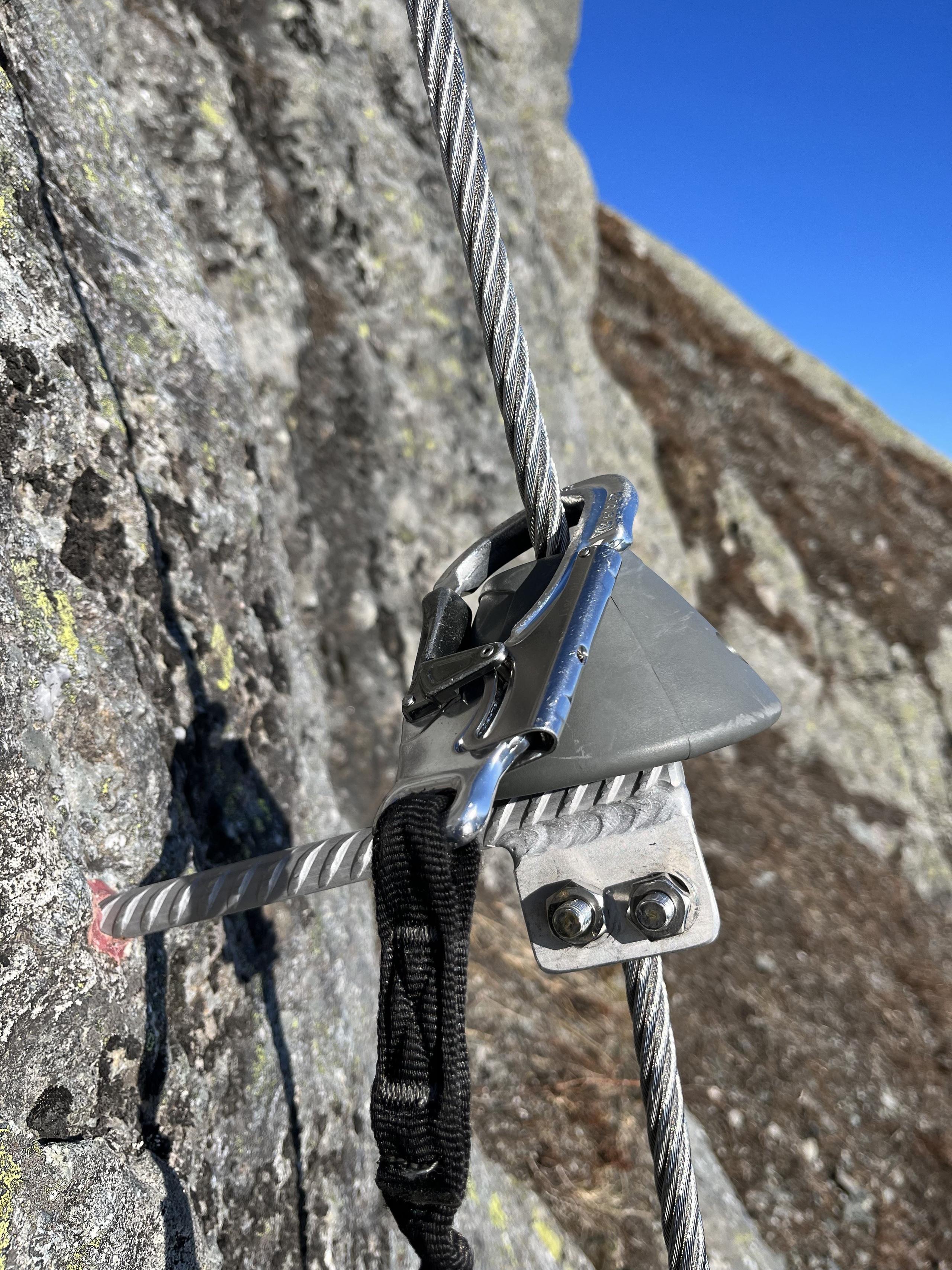 Close-up of a via ferrata steel cable clipped into a metal anchor on a rocky cliff under a clear blue sky.