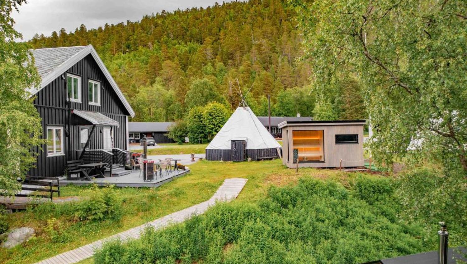 A scenic view of traditional black cabins, a tipi, and a modern structure amidst lush greenery.