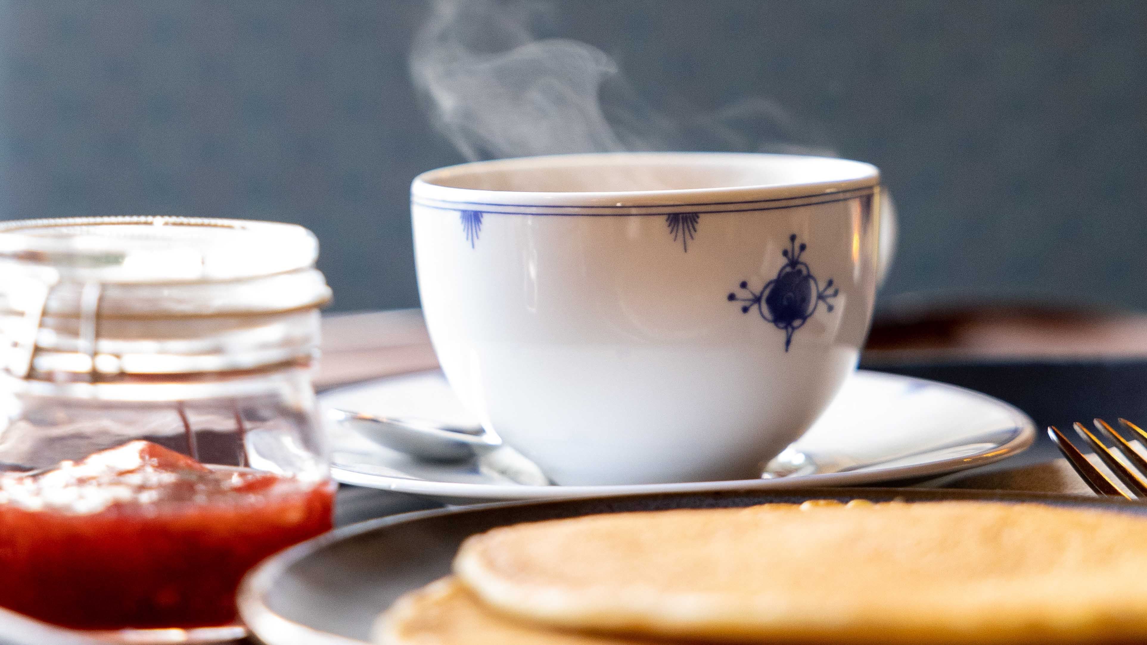 Wit keramisch kopje met blauw patroon op een schoteltje, stoom stijgt op, met een pot jam en brood erbij..