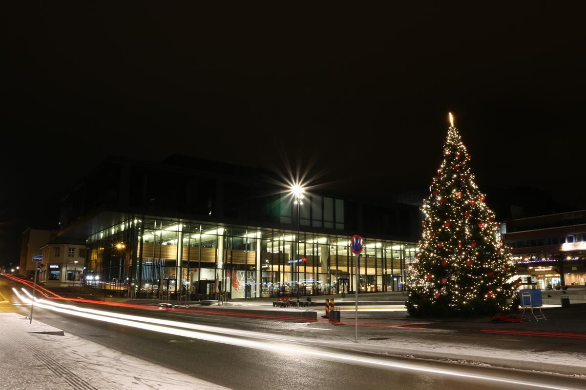 Et julepyntet torg foran Hamar kulturhus er avbildet om kvelden. En stor juletreinstallasjon med lys står til høyre, mens bygget bak er opplyst gjennom store glassfasader. Lysspor fra forbipasserende trafikk skaper bevegelse i forgrunnen.