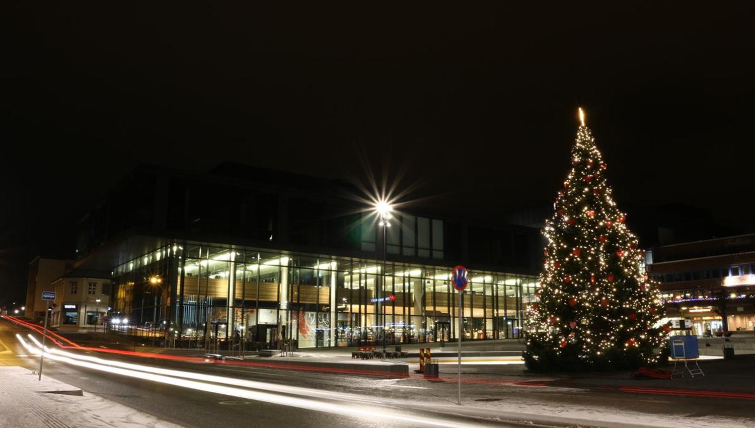 Et julepyntet torg foran Hamar kulturhus er avbildet om kvelden. En stor juletreinstallasjon med lys står til høyre, mens bygget bak er opplyst gjennom store glassfasader. Lysspor fra forbipasserende trafikk skaper bevegelse i forgrunnen.