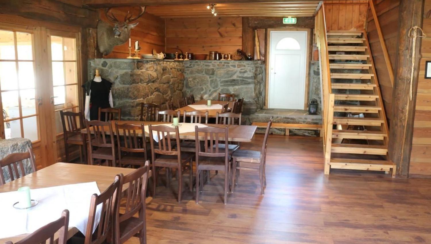 Wood-paneled lodge dining room with long tables and chairs, a stone wall with a mounted deer, and stairs by a white door, at Aabrekk Gard Trollbu in Oldedalen valley.