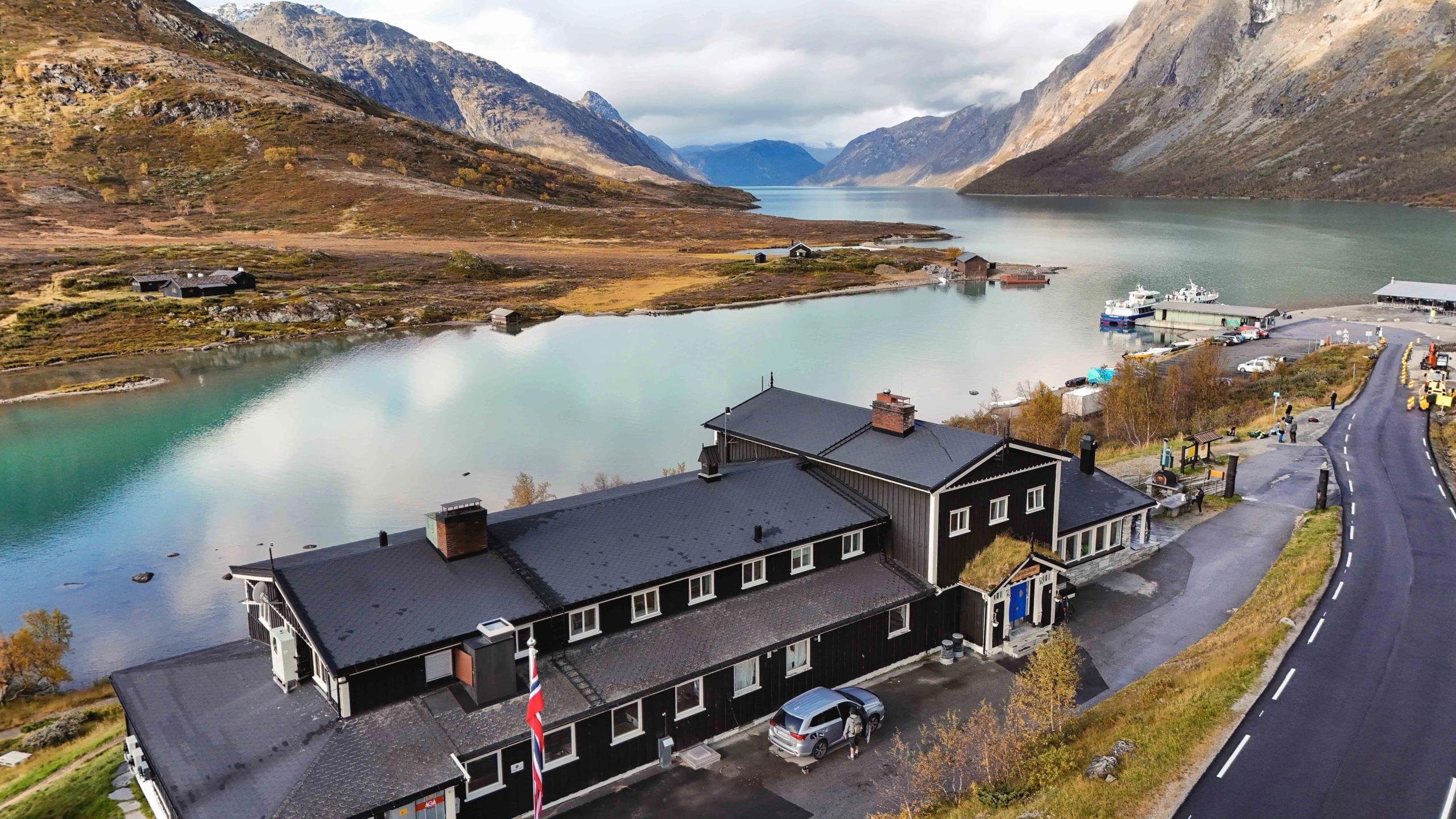 A black building beside a calm fjord with mountains in the background, small boats at a dock, and a road along the shore..