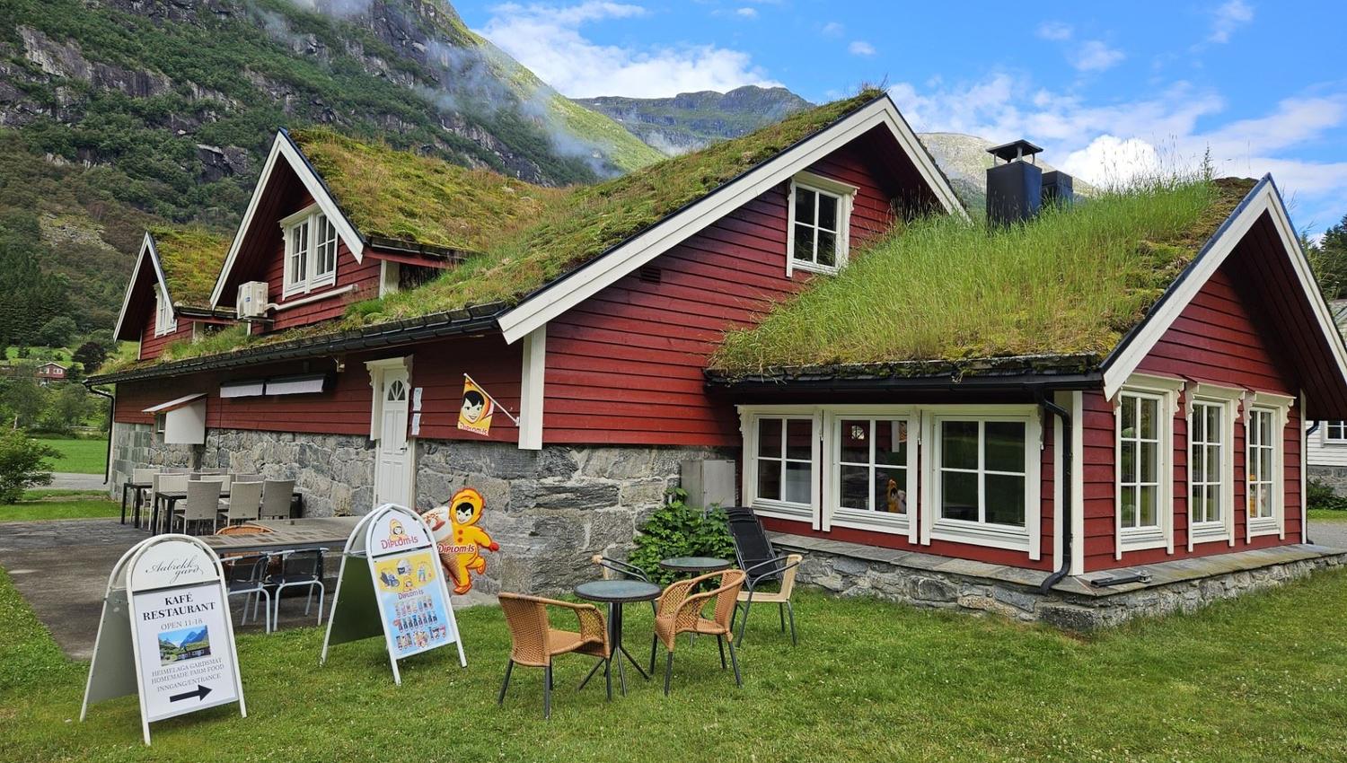 Red wooden cottage with a grass-covered roof, outdoor café tables and signs, set below green mountains under blue sky at Aabrekk Gard Trollbu in Oldedalen valley..