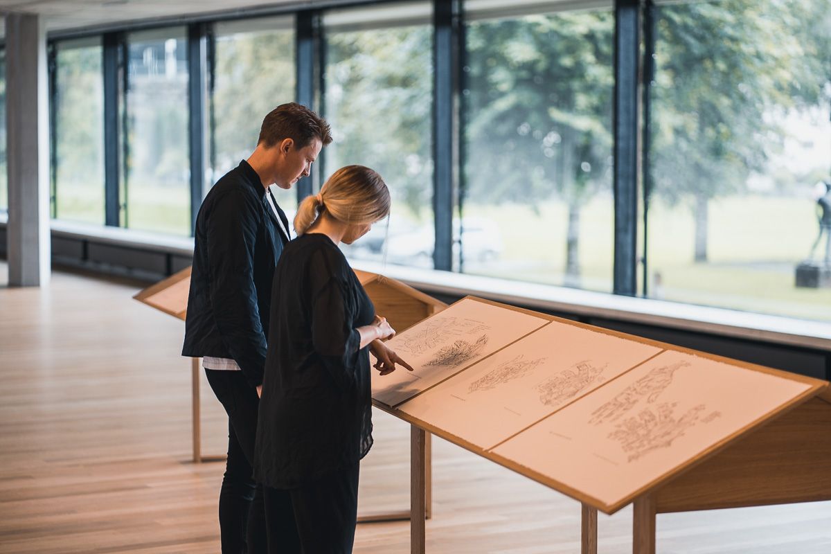 Visitors studying exhibition materials in the Kabuso Art House, with large windows overlooking nature.