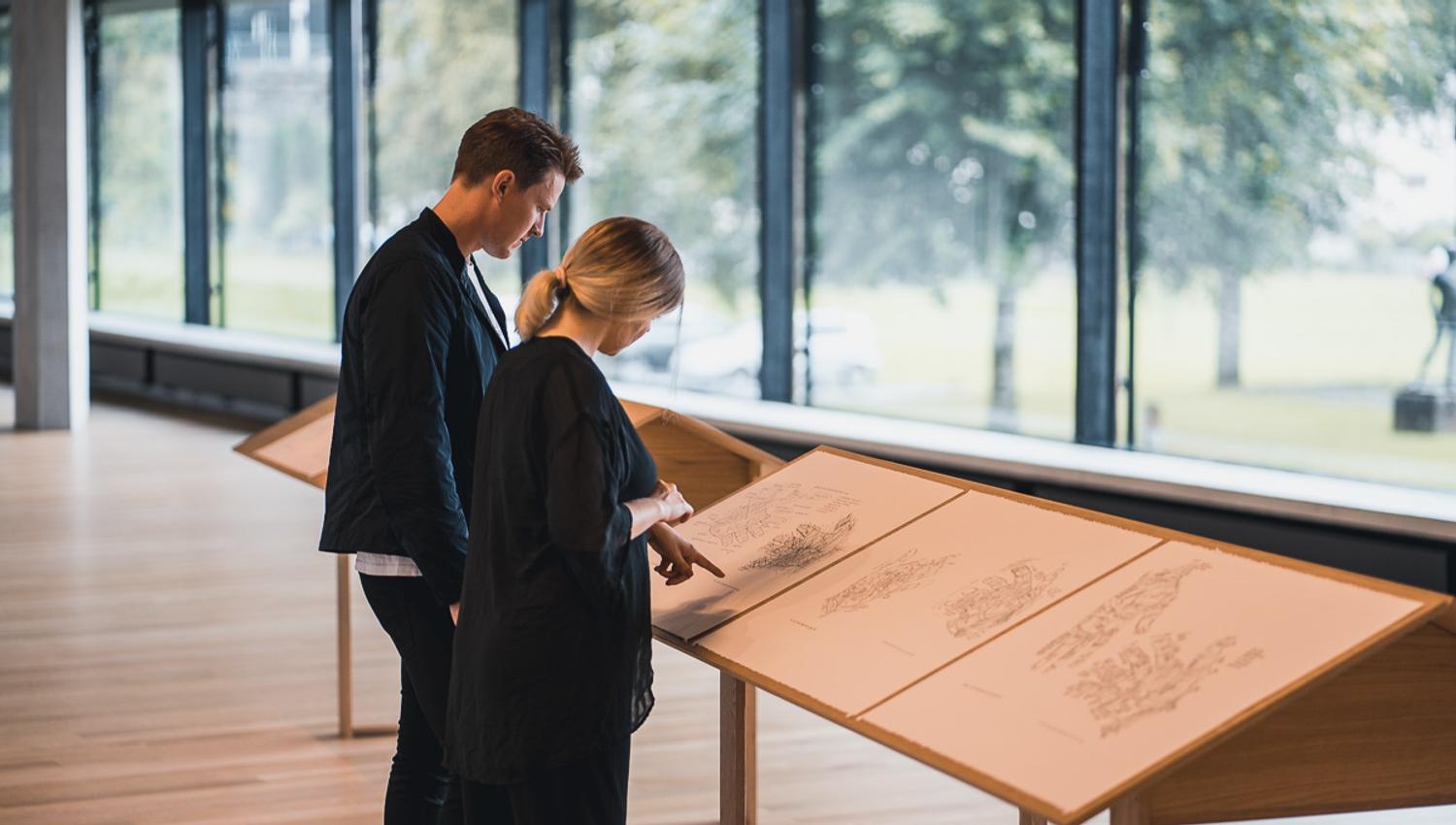 Visitors studying exhibition materials in the Kabuso Art House, with large windows overlooking nature.