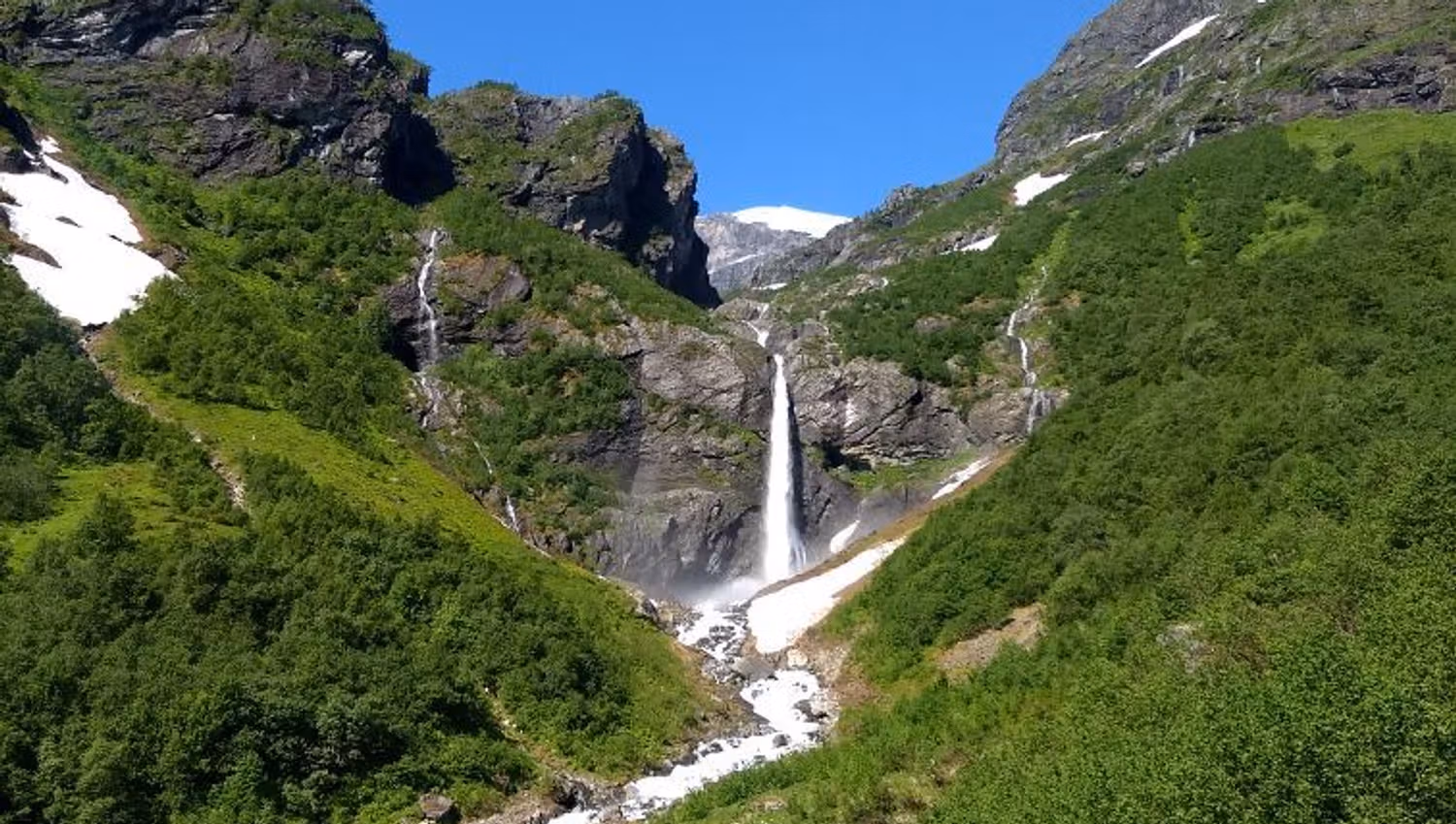 Tall waterfall drops between green mountain slopes, feeding a rocky stream with snow patches under a blue sky.