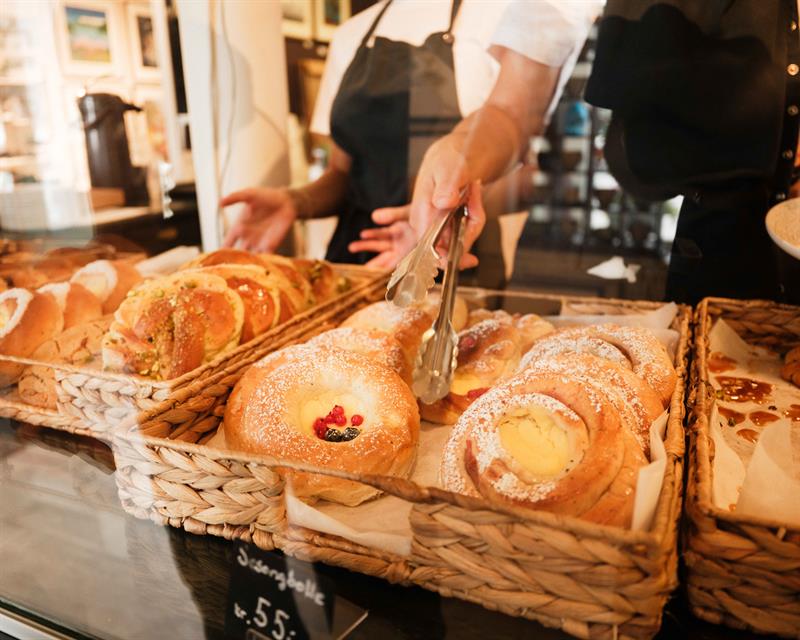 Freshly baked pastries are displayed in the counter inside Iversen Bakery and Gallery, while a staff member uses tongs to place baked goods behind the