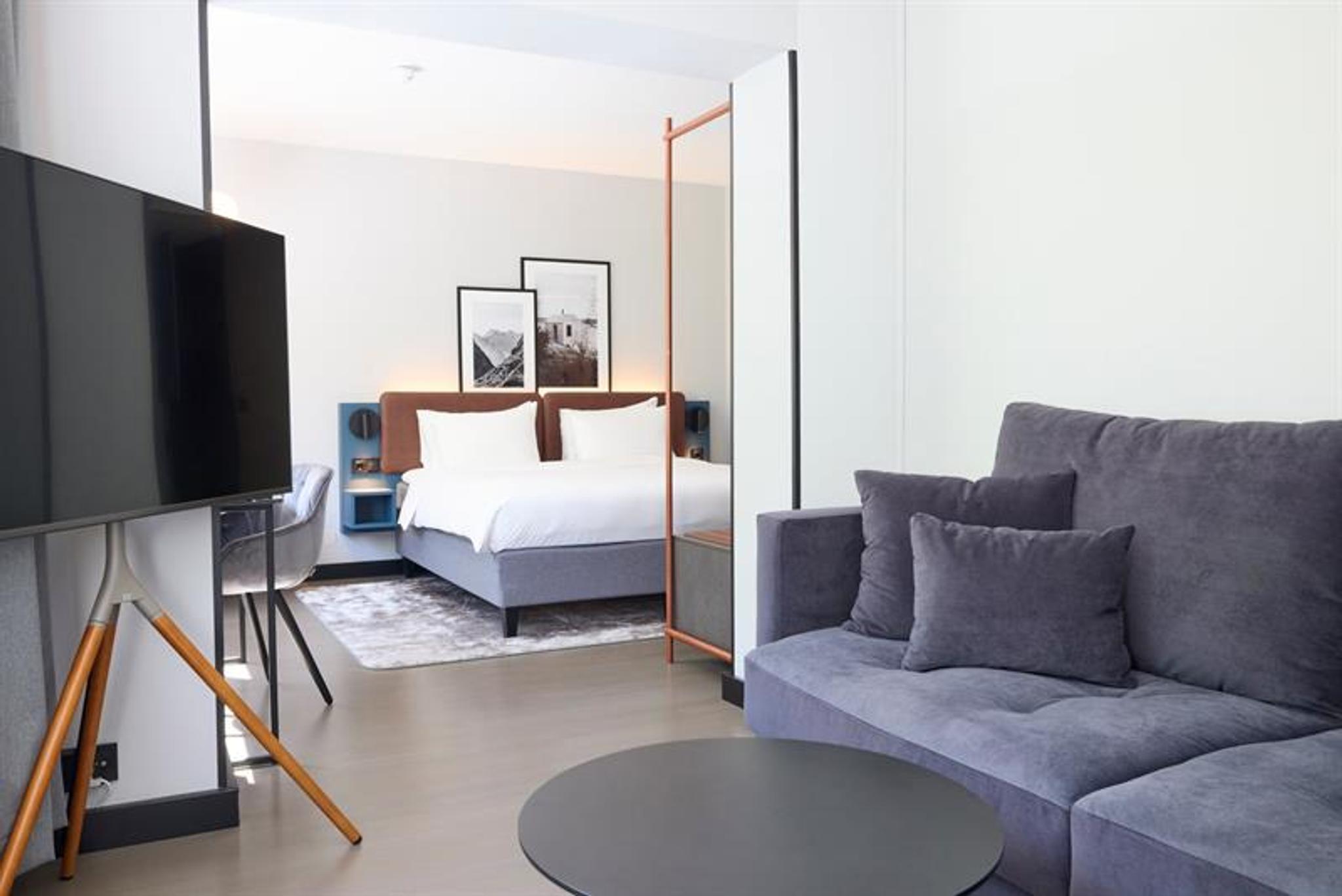 Moderne hotellrom med dobbeltseng, sofa, skrivebord og TV i lyse, stilrene omgivelser.