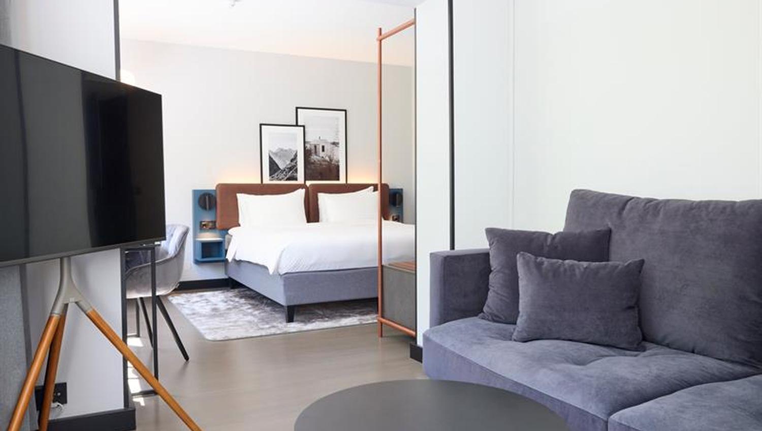 Moderne hotellrom med dobbeltseng, sofa, skrivebord og TV i lyse, stilrene omgivelser.