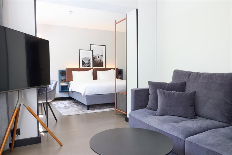 Moderne hotellrom med dobbeltseng, sofa, skrivebord og TV i lyse, stilrene omgivelser.