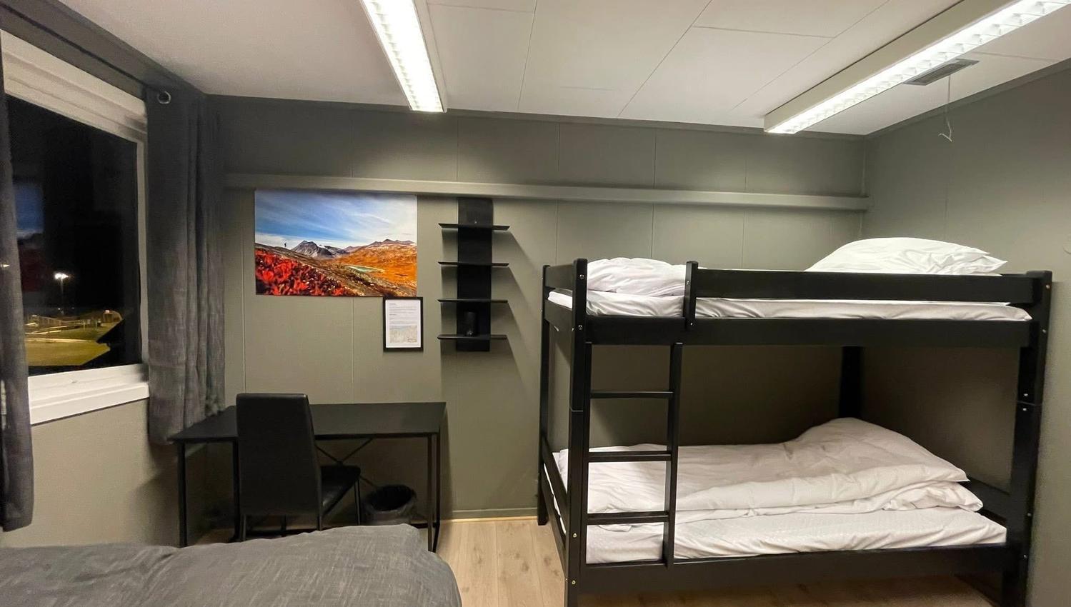 Interior of a small room with a black bunk bed, a desk and chair by a window, and wall shelves.