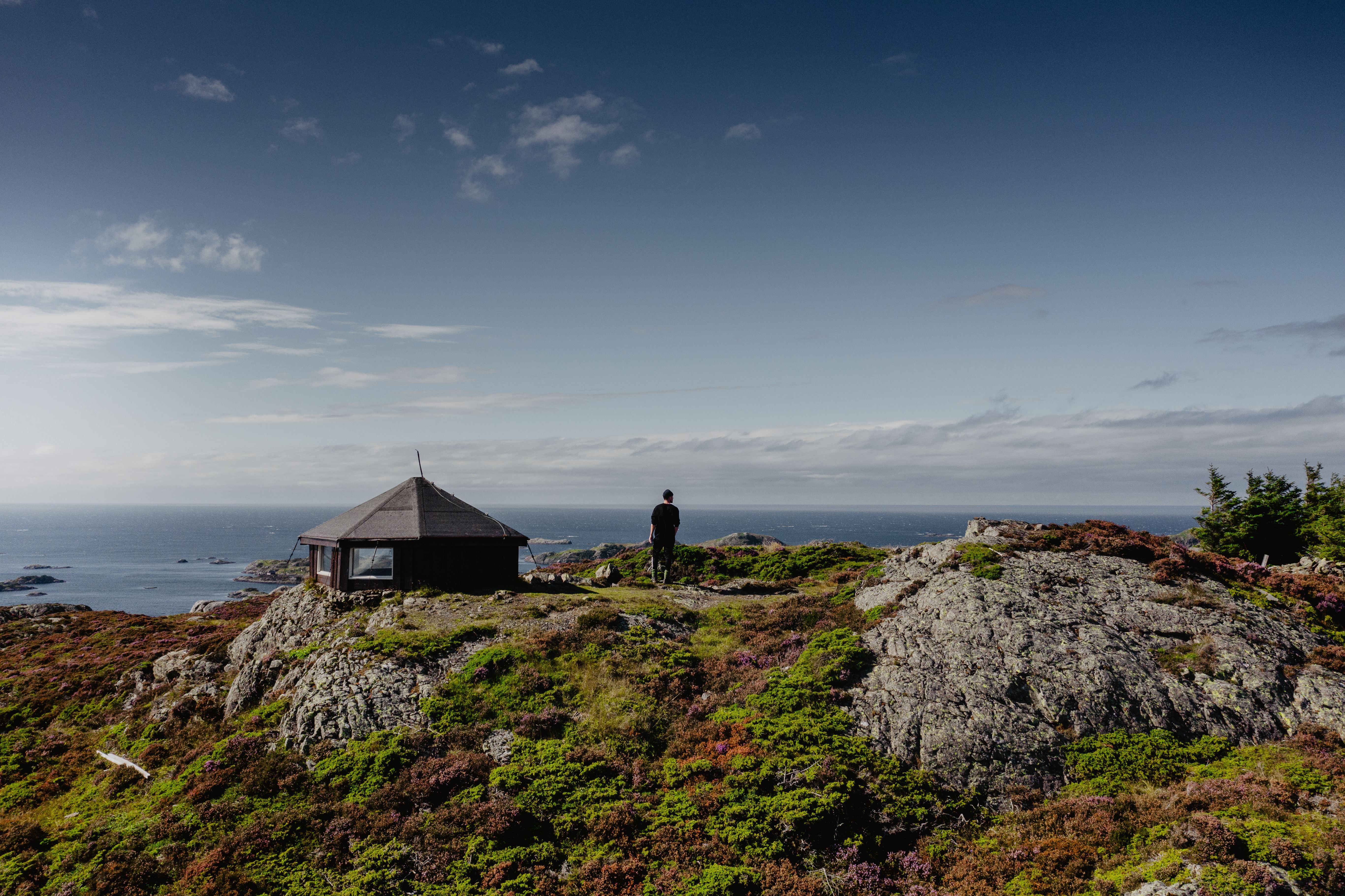 A lone person stands on rocky coastal hills near a small cabin, overlooking the ocean under a blue sky.