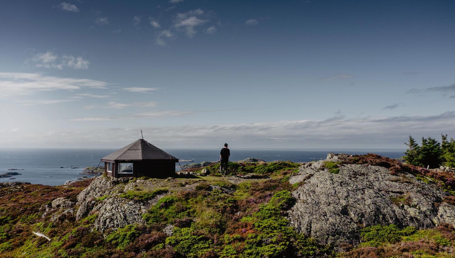A lone person stands on rocky coastal hills near a small cabin, overlooking the ocean under a blue sky.