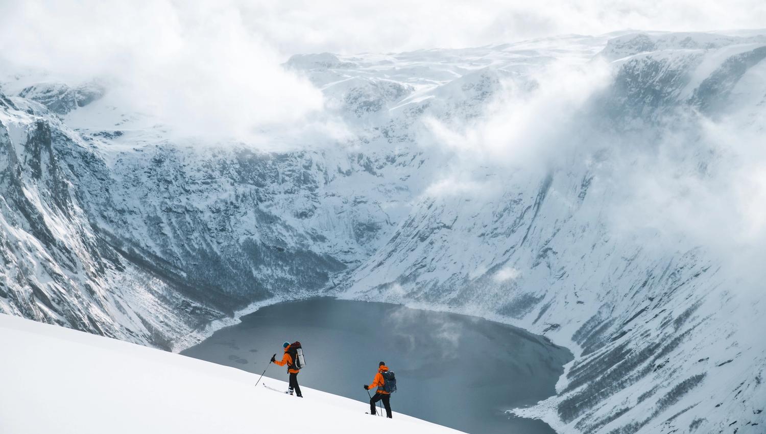 Two people skiing beside a lake surrounded by snowy mountains.