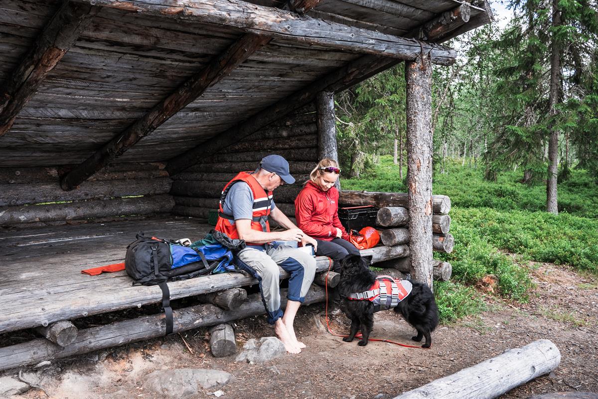 A man and woman in life jackets sitting under a wooden shelter with a black dog on a leash.