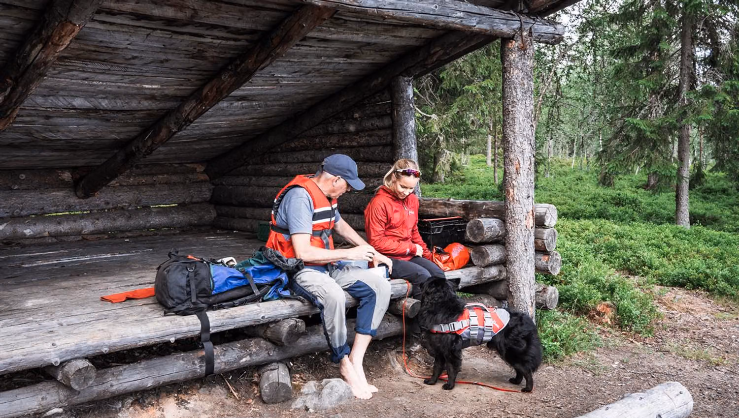 A man and woman in life jackets sitting under a wooden shelter with a black dog on a leash.