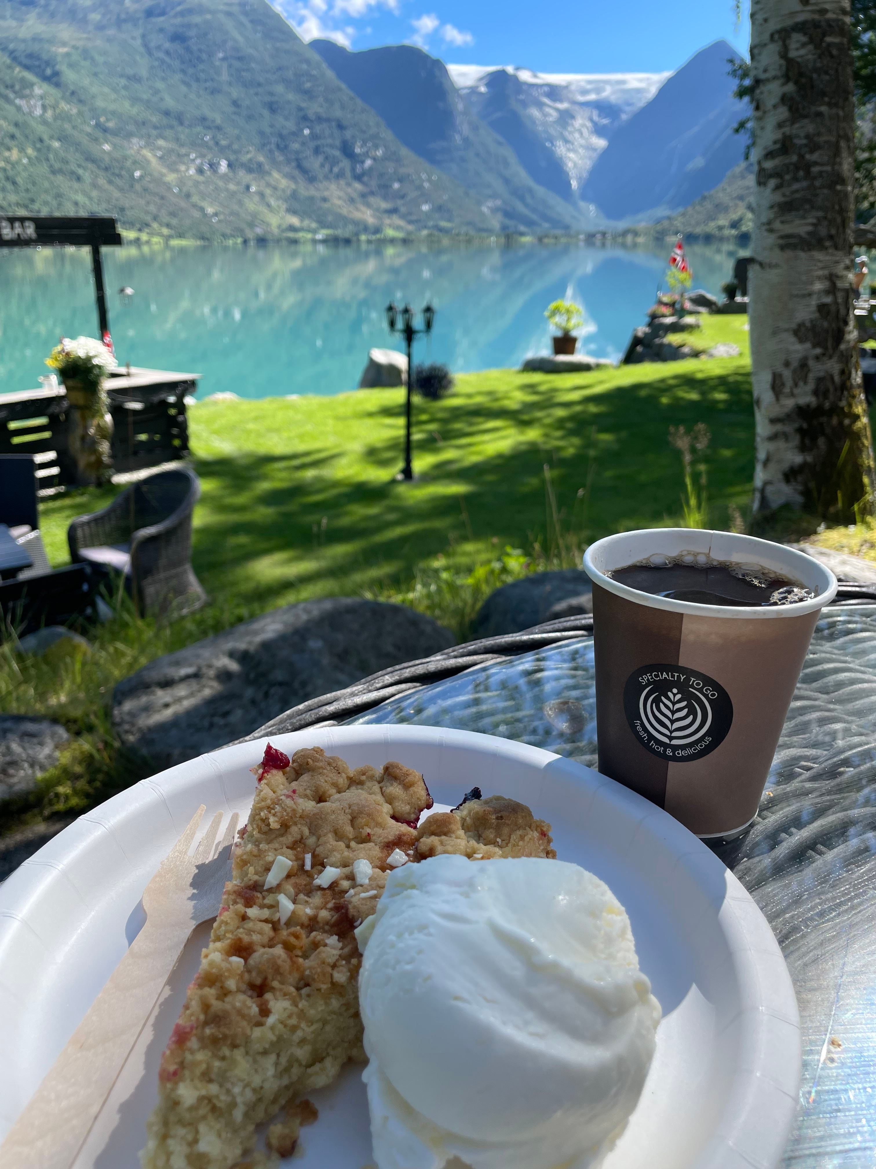 Paper cup of coffee and a slice of crumb cake with vanilla ice cream on a table overlooking a calm fjord and mountains.
