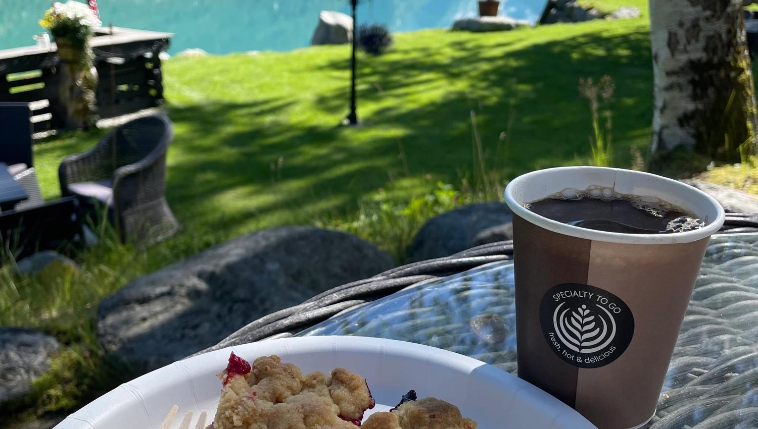 Paper cup of coffee and a slice of crumb cake with vanilla ice cream on a table overlooking a calm fjord and mountains.