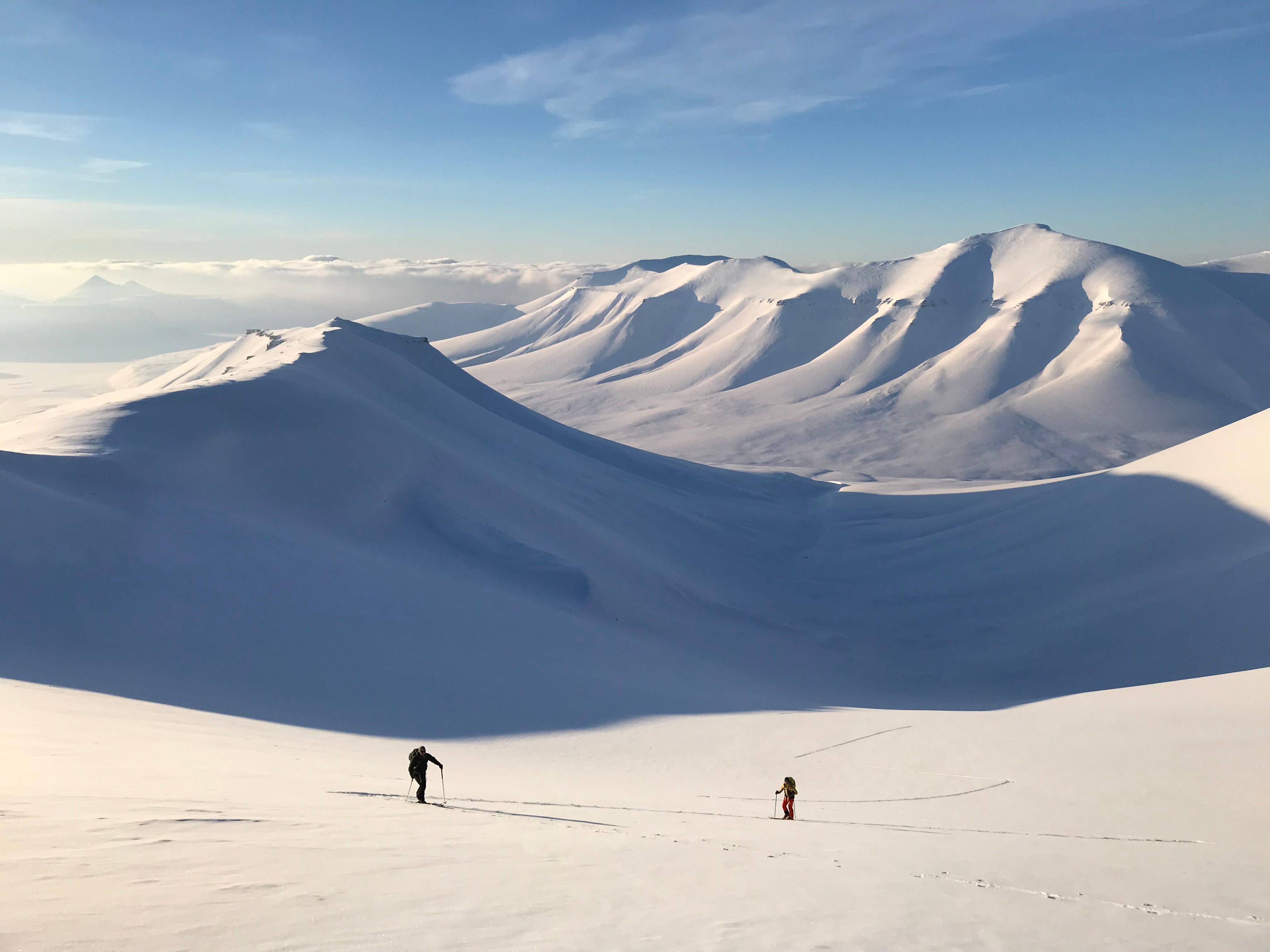 Two persons skiing up a mountain with a long valley and a mountainous landscape in the background