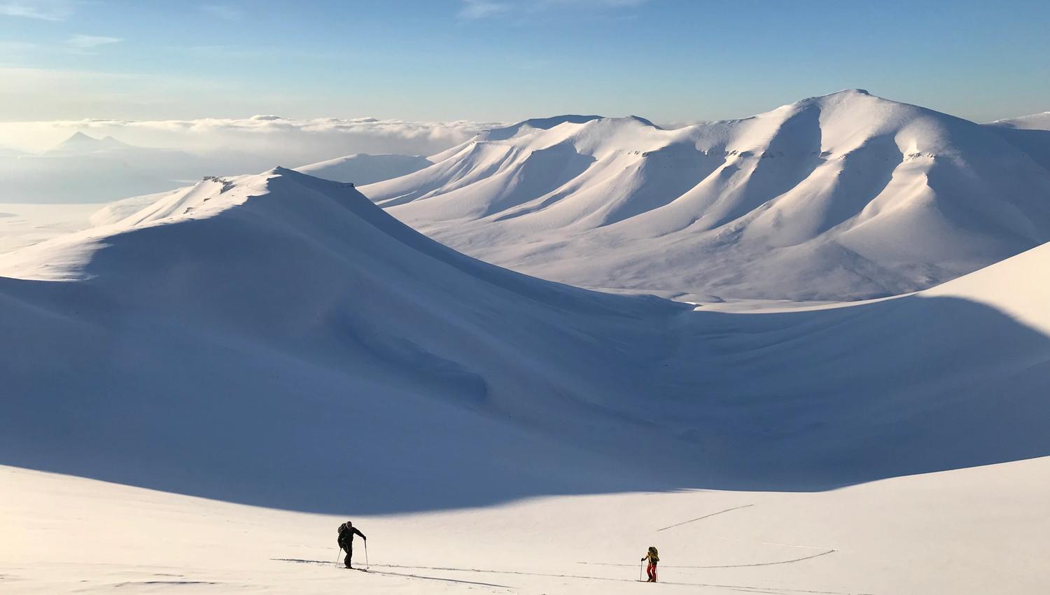 Two persons skiing up a mountain with a long valley and a mountainous landscape in the background