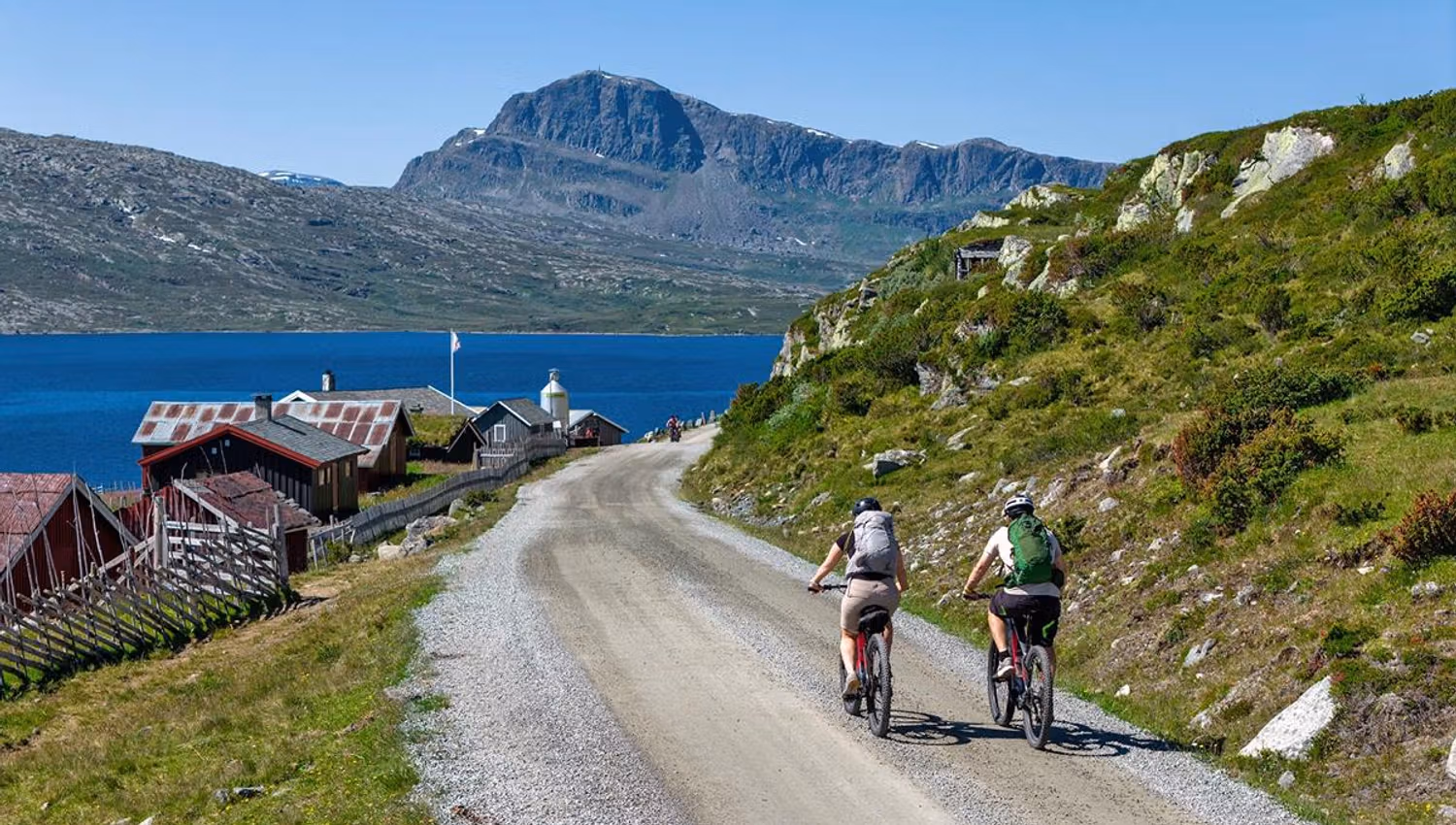 To syklister på grusvei langs vann med fjell i bakgrunnen.