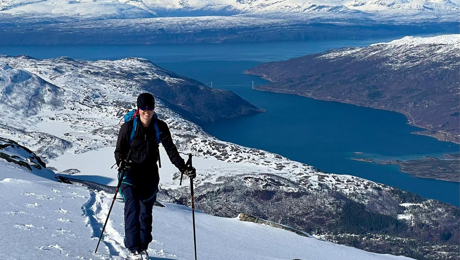 A person on backcountry skis climbs a snowy ridge with poles, overlooking a blue fjord and distant peaks.