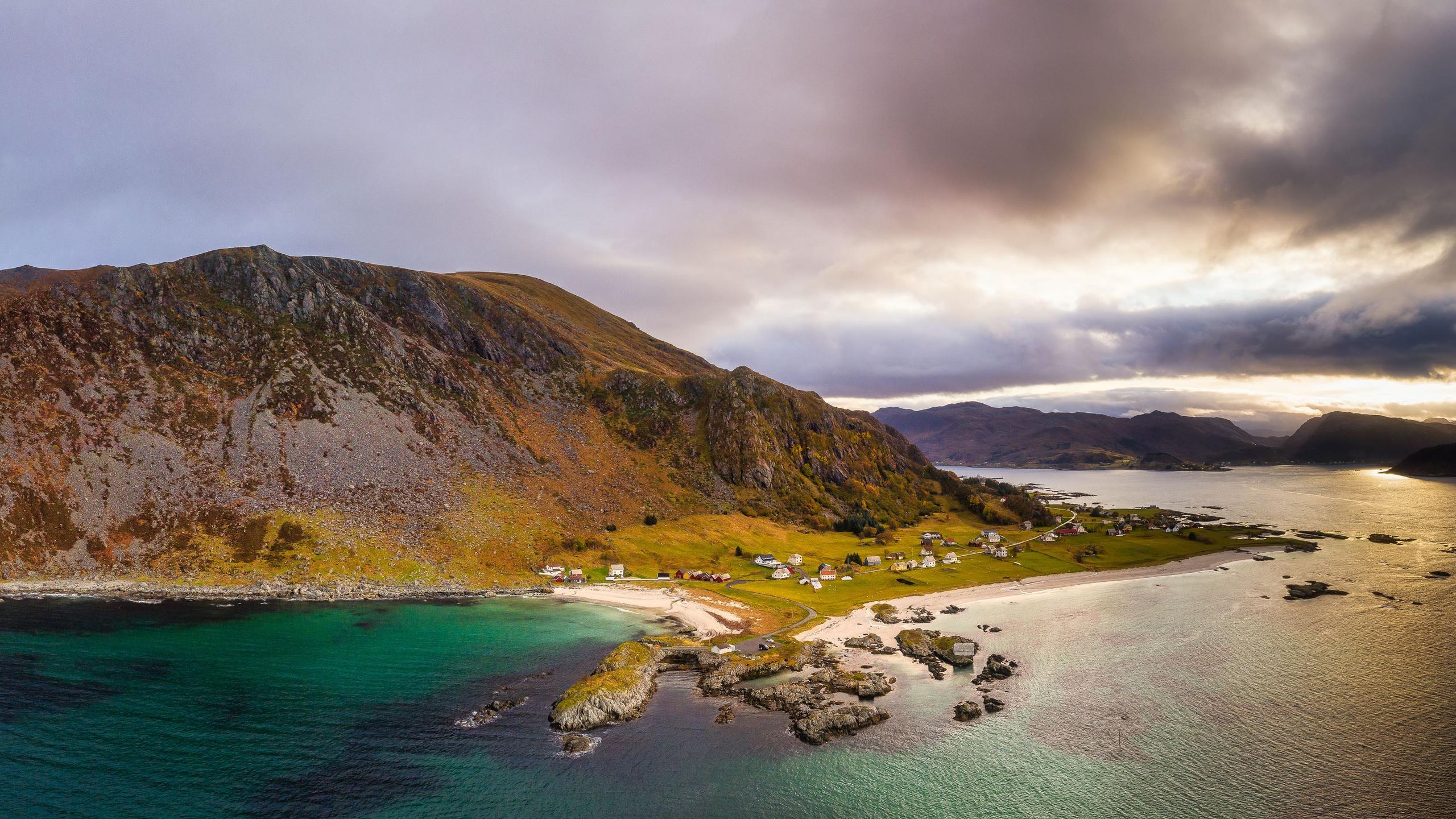 Grotlesanden. A rugged mountain ridge overlooks a small coastal village, sandy beach, rocky shore, and calm sea under cloudy sunset light..