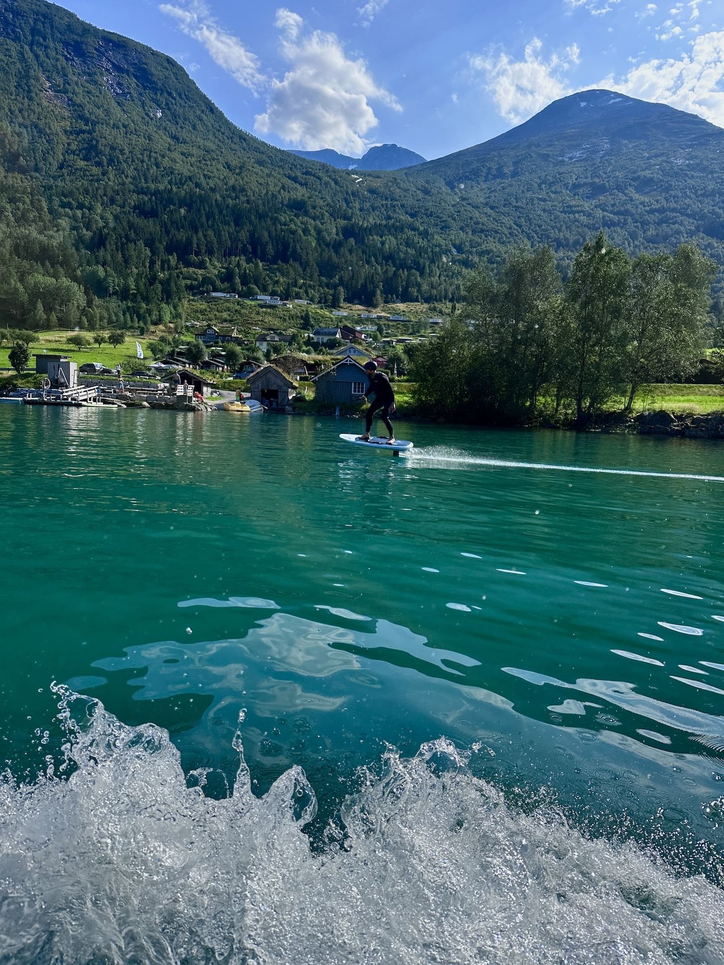 Person riding a hydrofoil board on turquoise lake water, with green mountains, lakeside cabins, and clouds..