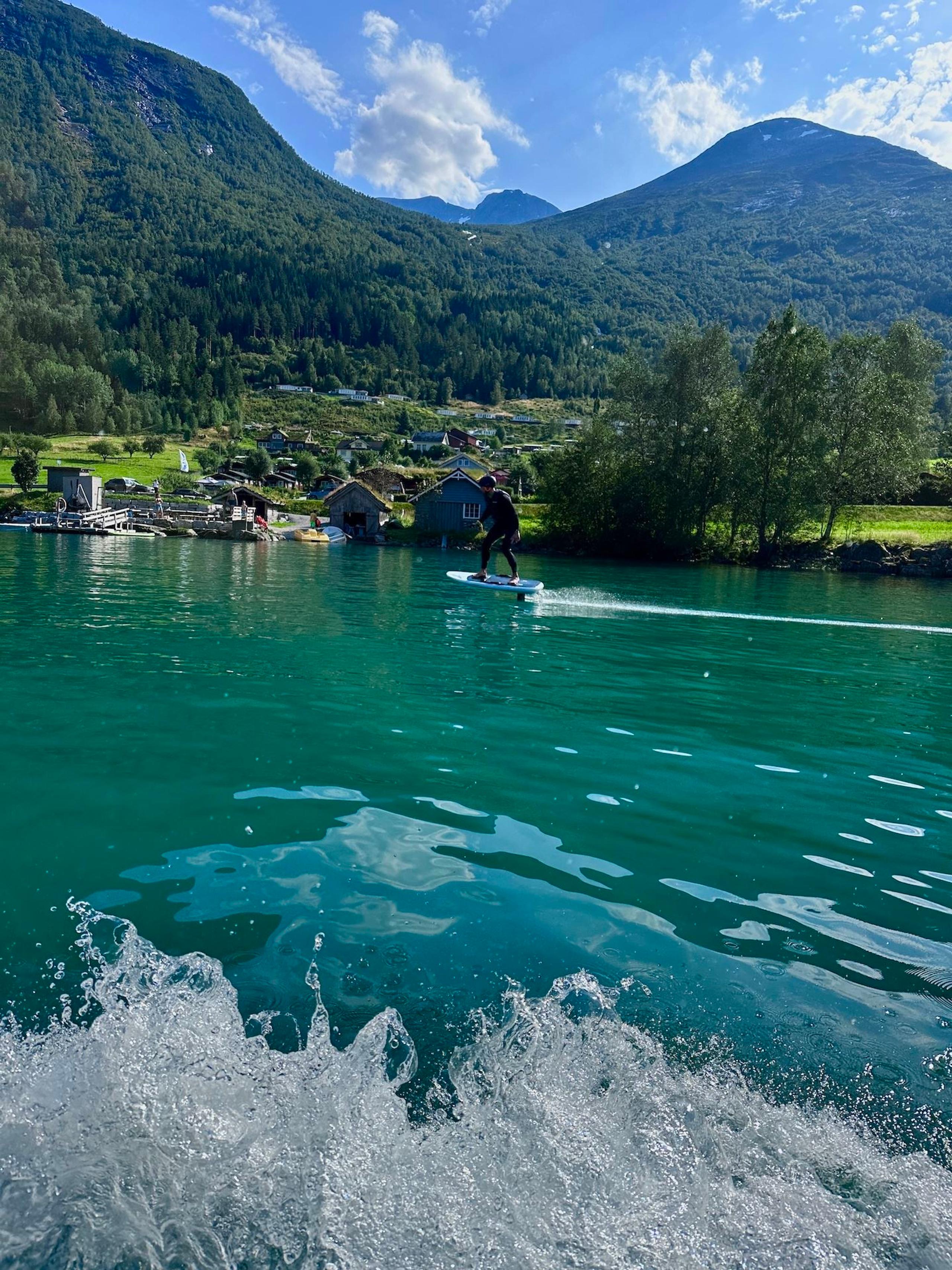 Person riding a hydrofoil board on turquoise lake water, with green mountains, lakeside cabins, and clouds..