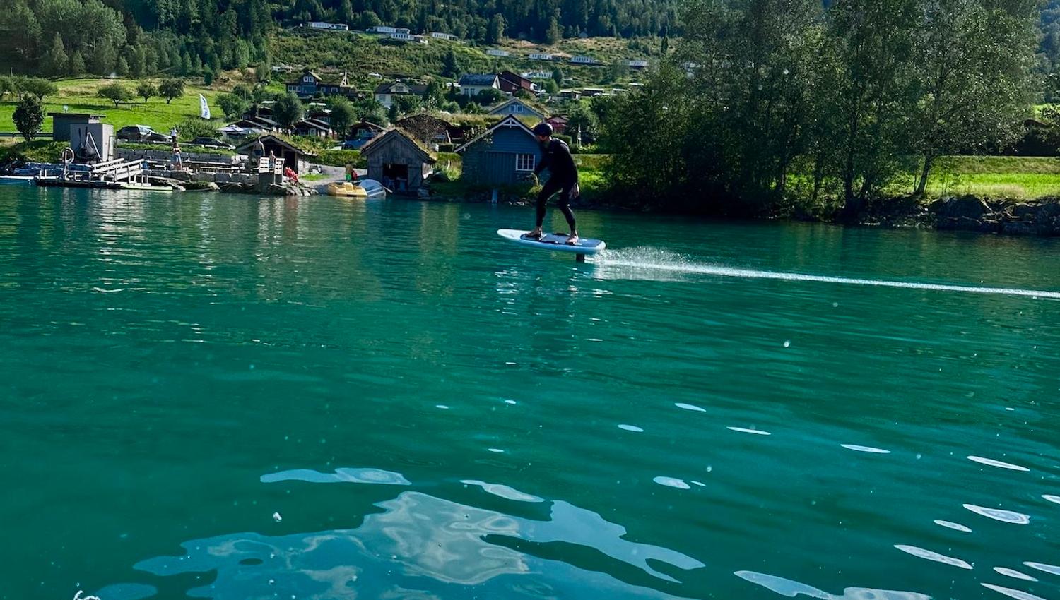 Person riding a hydrofoil board on turquoise lake water, with green mountains, lakeside cabins, and clouds..