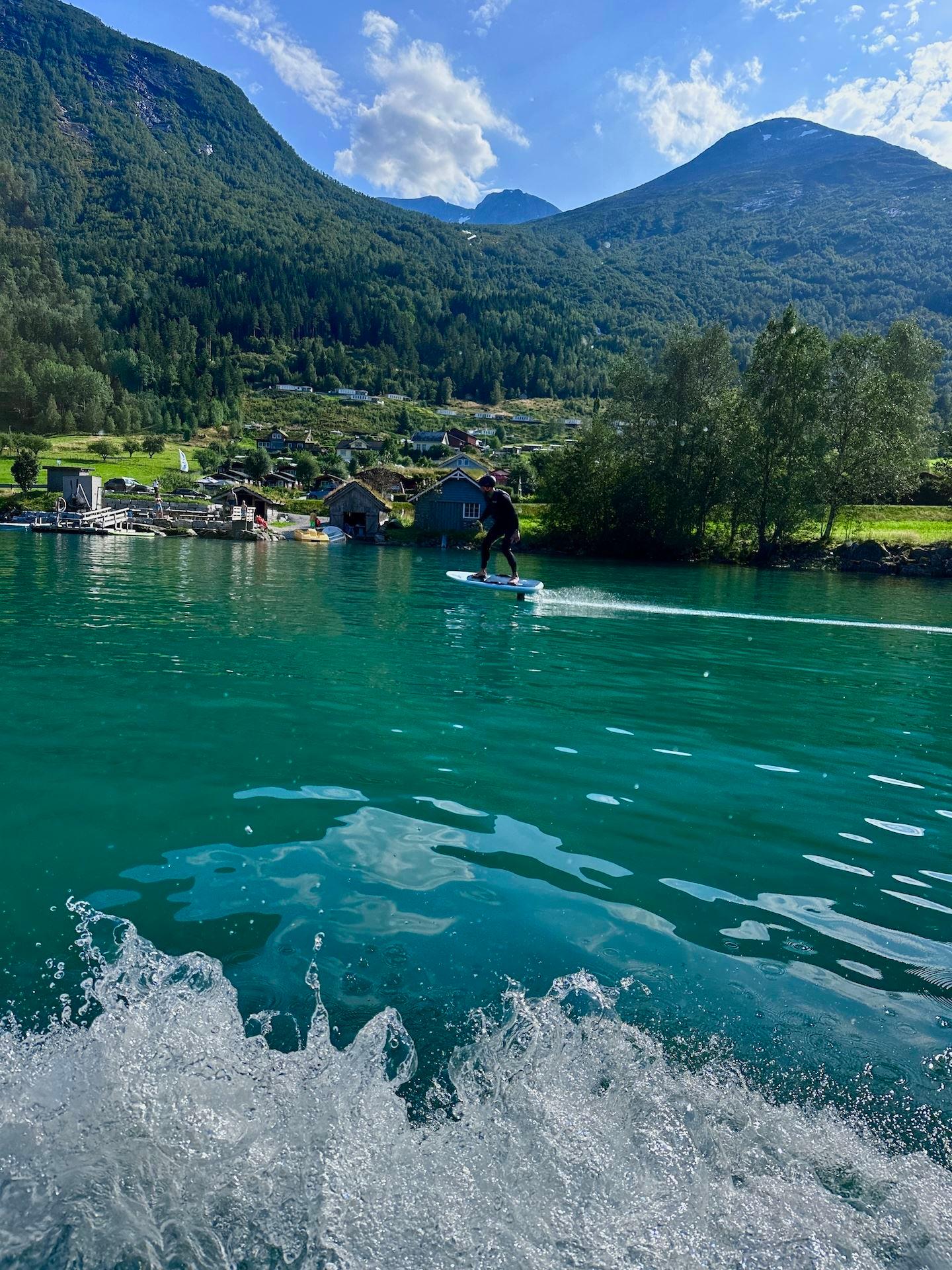 Person riding a hydrofoil board on turquoise lake water, with green mountains, lakeside cabins, and clouds..