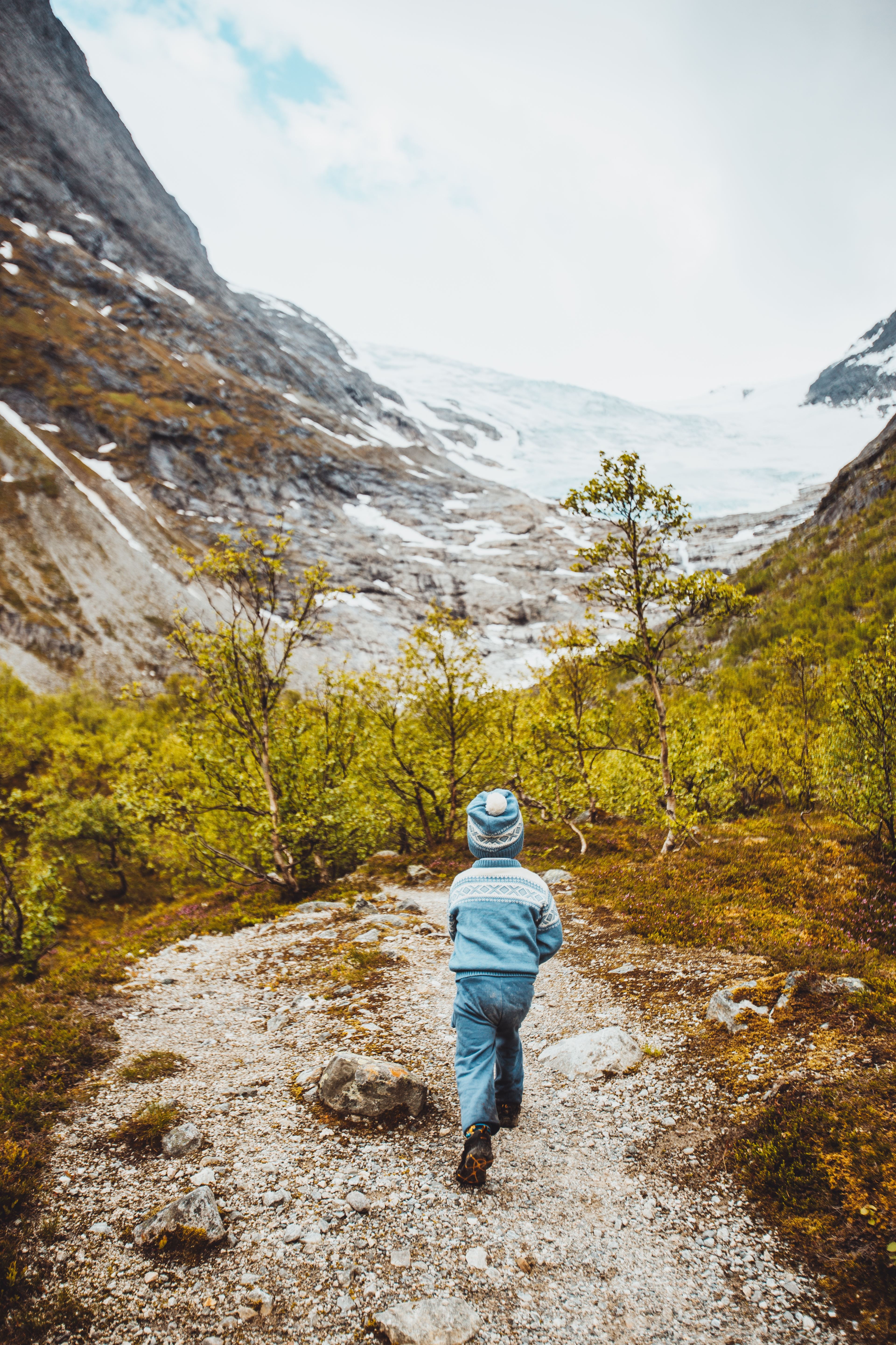 A child in a blue knit hat and sweater walks on a rocky trail through shrubs toward snowy mountains and a glacier.