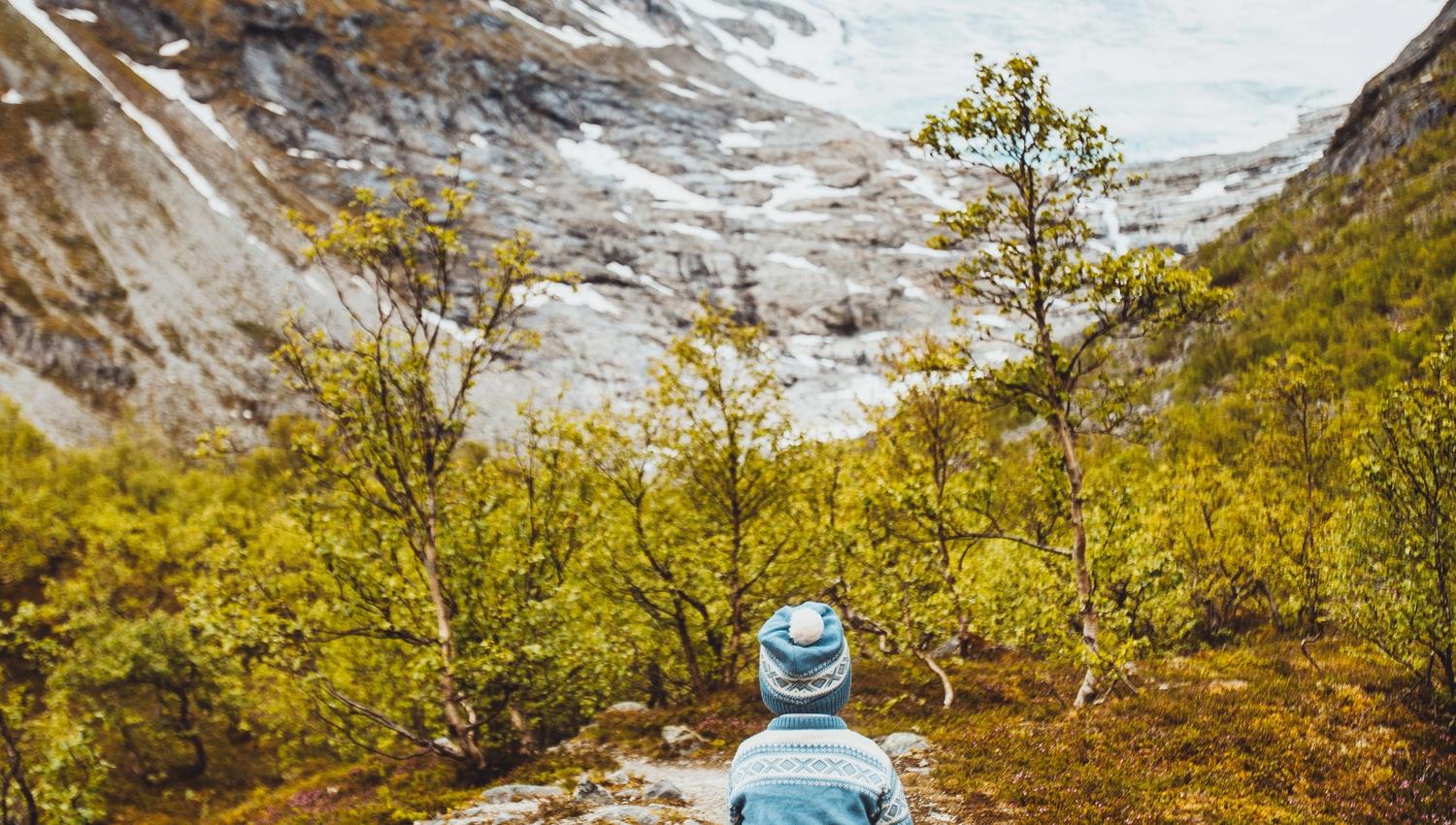 A child in a blue knit hat and sweater walks on a rocky trail through shrubs toward snowy mountains and a glacier.