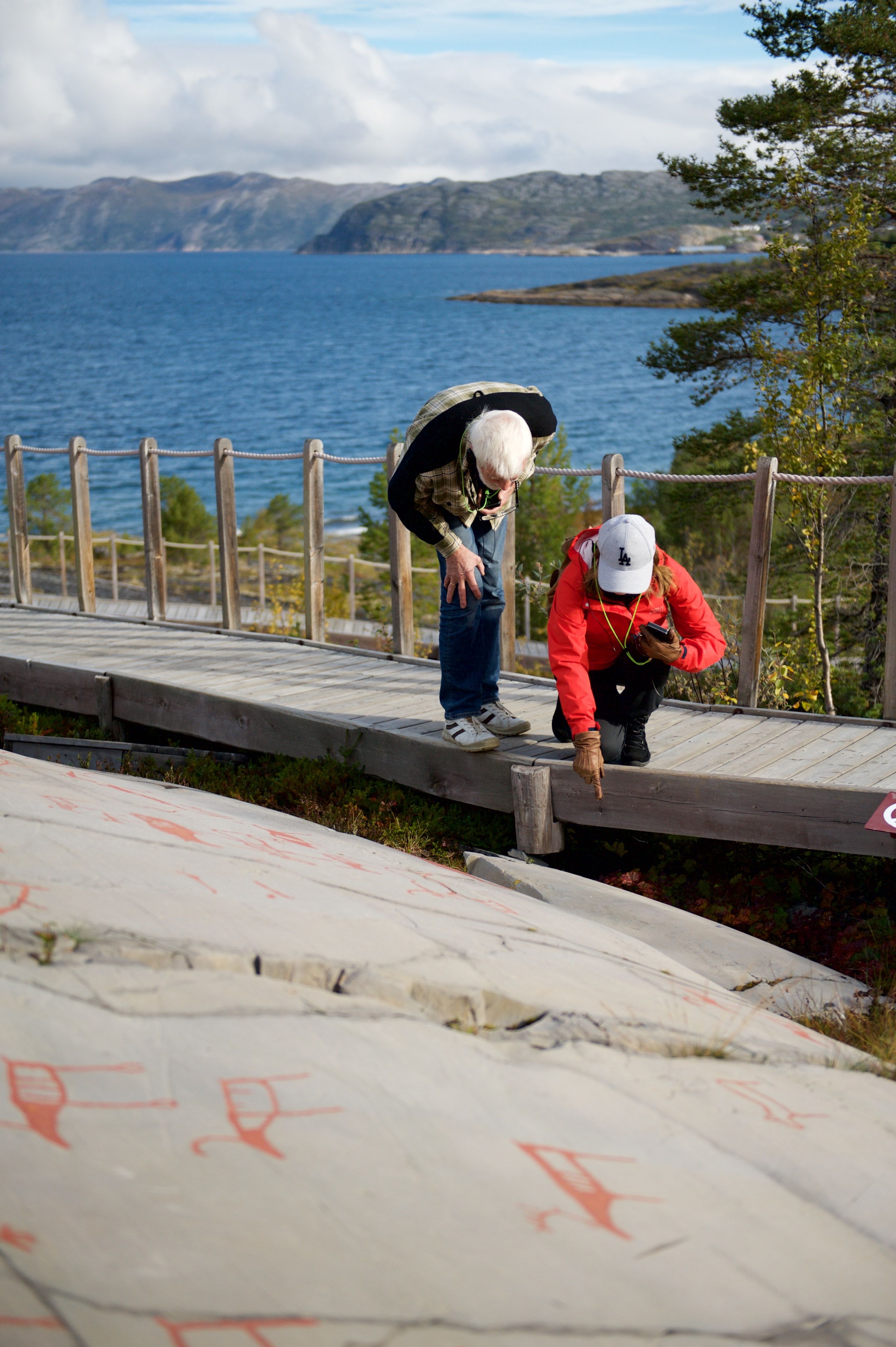 Explore the intriguing world of ancient art as two visitors inspect petroglyphs along a scenic coastal walkway. The backdrop features stunning cliffs and serene waters, making this a perfect spot for history enthusiasts and nature lovers alike.