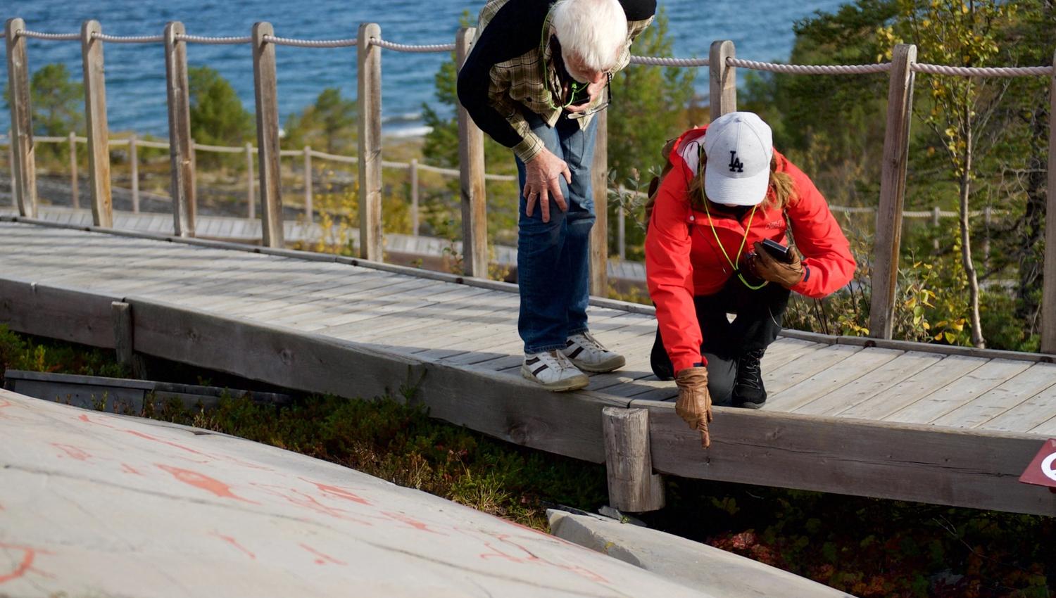 Explore the intriguing world of ancient art as two visitors inspect petroglyphs along a scenic coastal walkway. The backdrop features stunning cliffs and serene waters, making this a perfect spot for history enthusiasts and nature lovers alike.